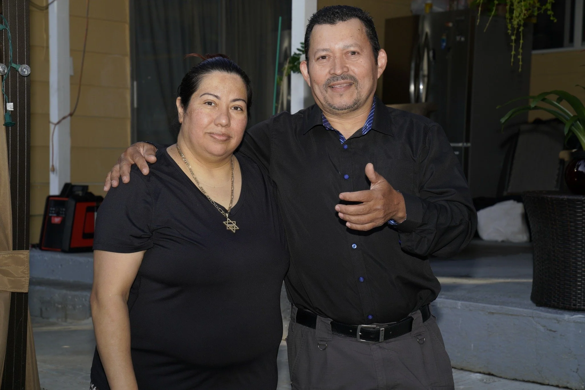 A man and woman standing close together, with the man's arm around the woman's shoulder, both smiling slightly at the camera. They are dressed in black shirts, and are inside a room with yellow walls and some household items in the background.