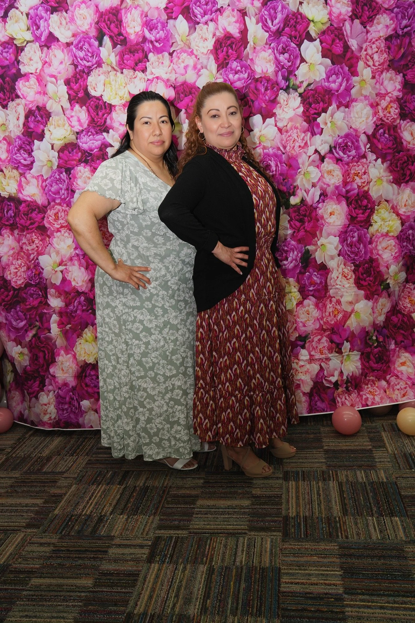 Two women standing in front of a vibrant floral backdrop with pink, purple, and white flowers. One woman is dressed in a light green floral dress, and the other in a patterned dress with a black blazer. Both are posing with their hands on their hips.