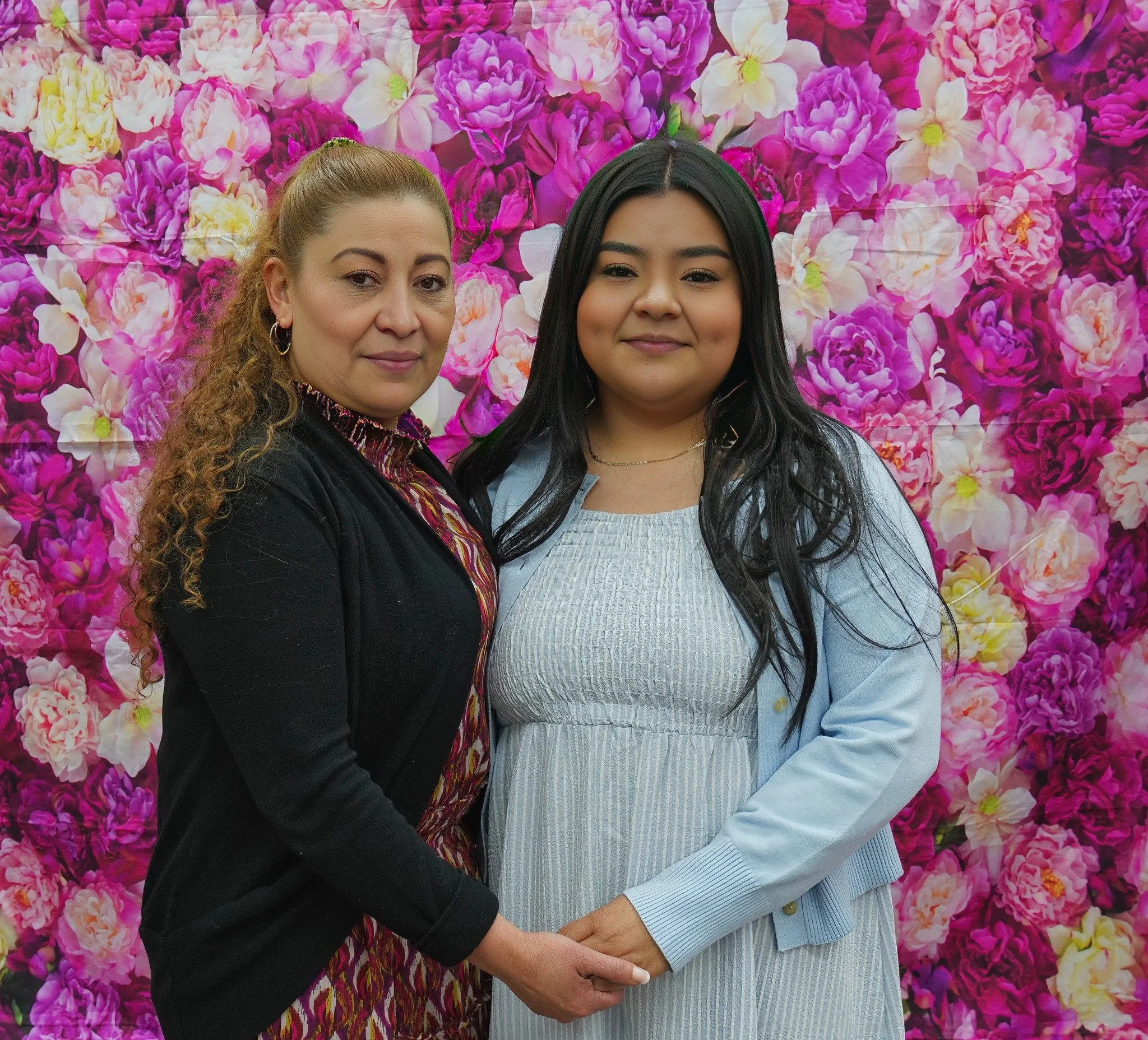 Two women standing close together in front of a pink floral backdrop, holding hands and smiling.