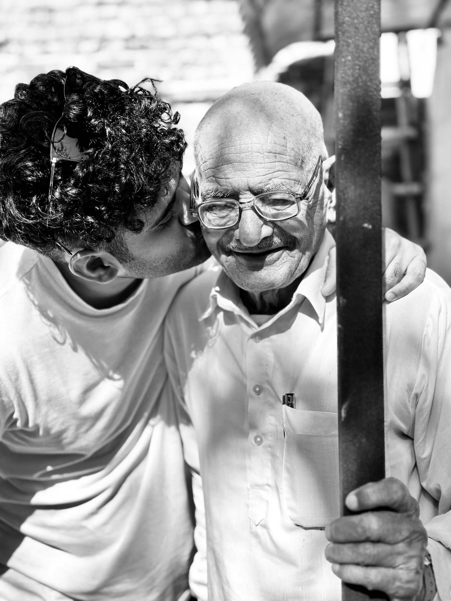 A young man kisses an elderly man on the cheek, both smiling, with the elderly man holding a vertical pole.