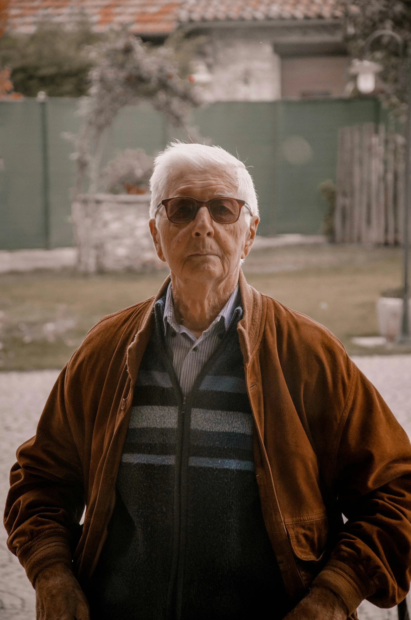 An elderly man with white hair wearing sunglasses, a striped button-up shirt, a zip-up sweater, and a brown jacket standing outside.