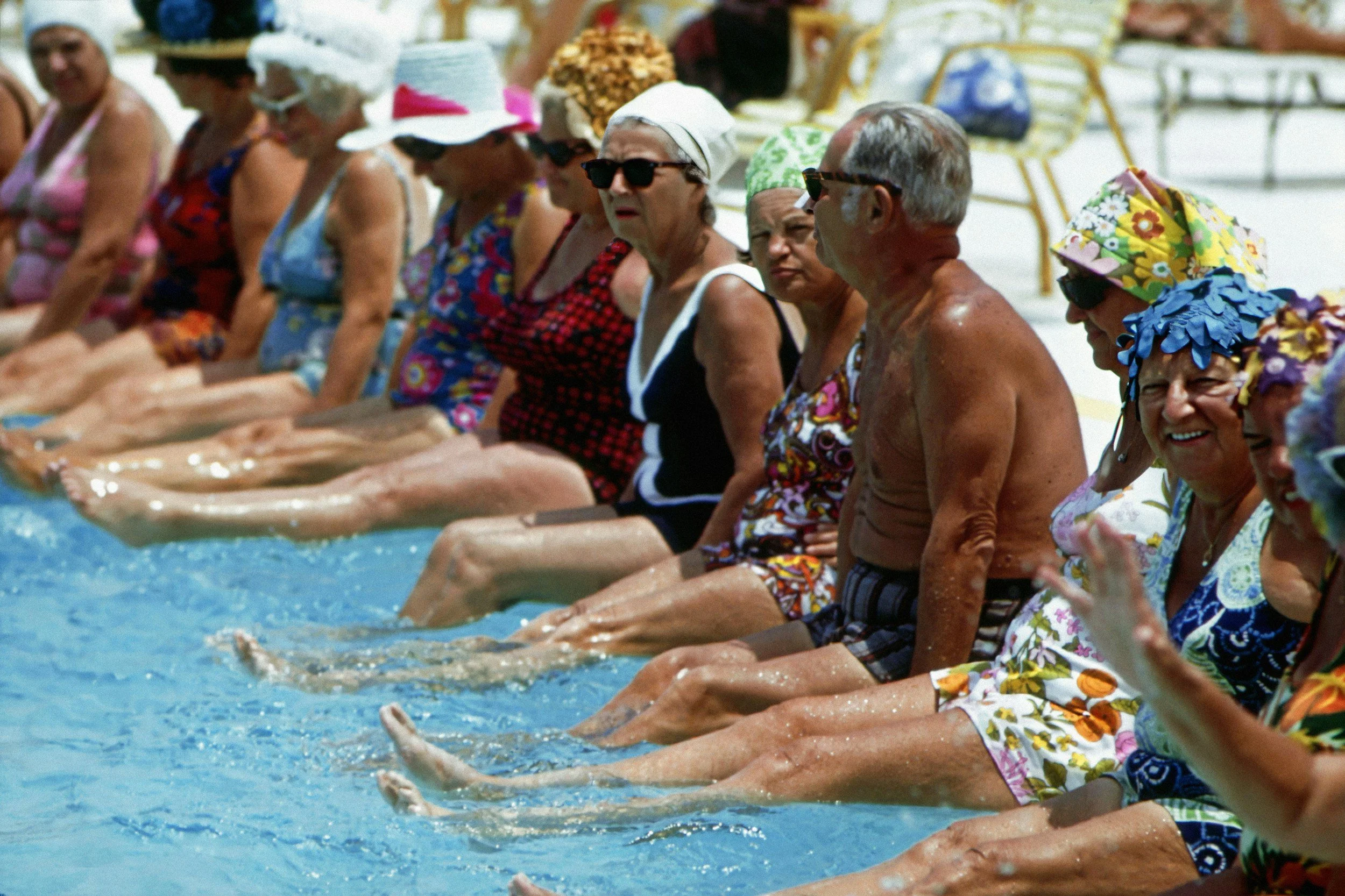 Senior group of women and men sitting by poolside with their legs in the water, wearing colorful swimsuits, hats, and sunglasses, on a bright sunny day.