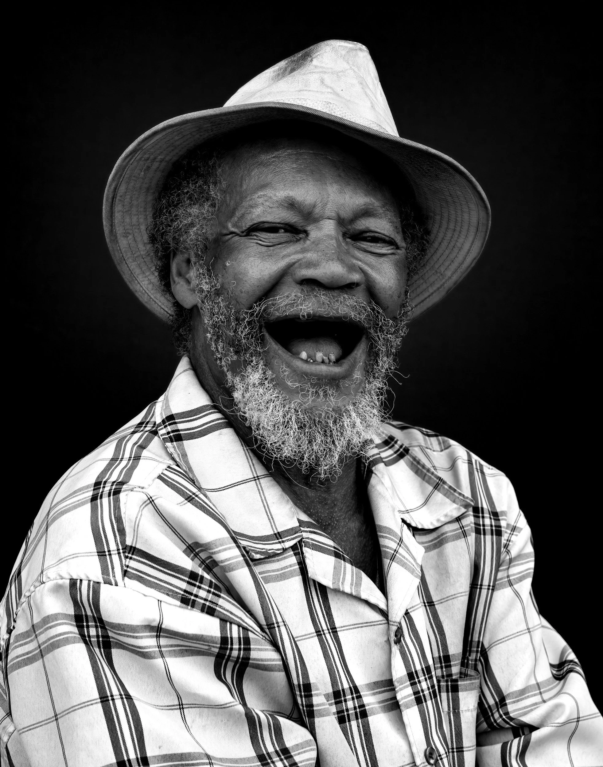 Black and white portrait of an elderly man with a beard, smiling widely with missing teeth, wearing a cowboy hat and checked shirt.