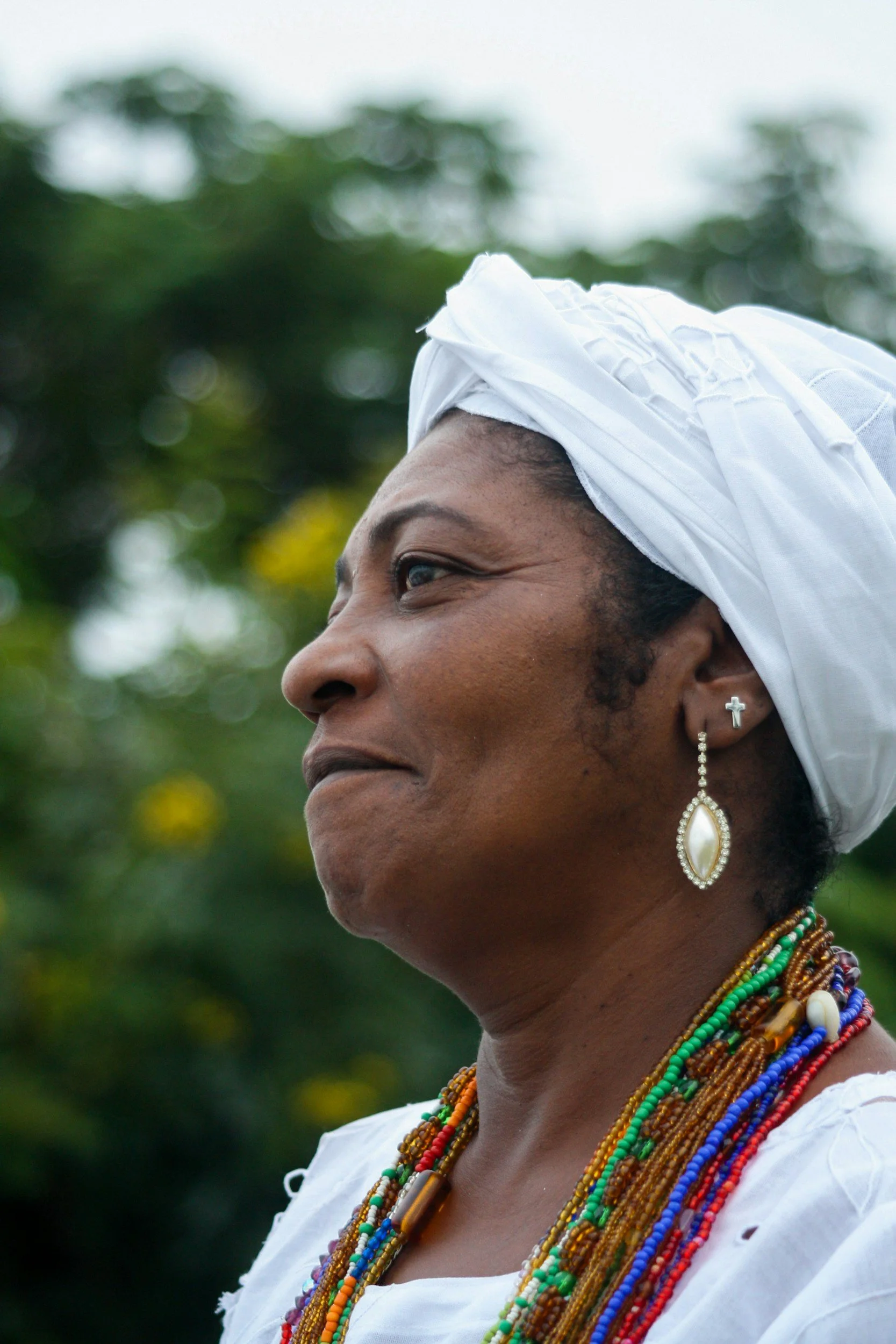 Profile of a woman wearing a white headwrap, white dress, and multiple colorful beaded necklaces, earrings, and a small cross earring, outdoors with greenery in the background.