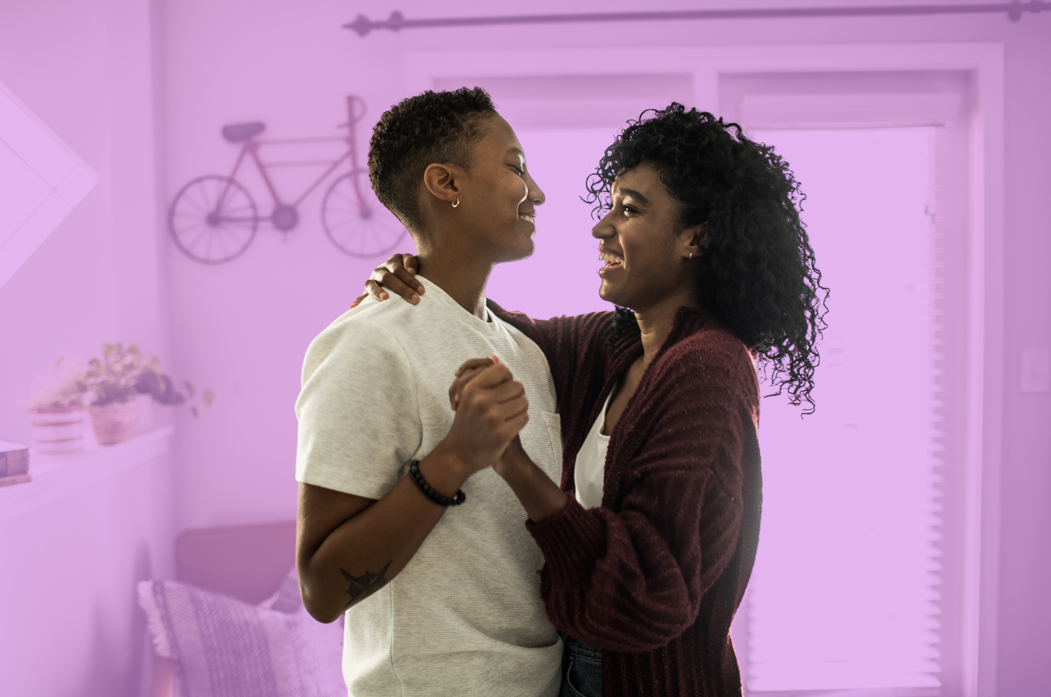 Two women smiling, holding hands, dancing together in a brightly lit room with purple walls and a bicycle wall decoration.