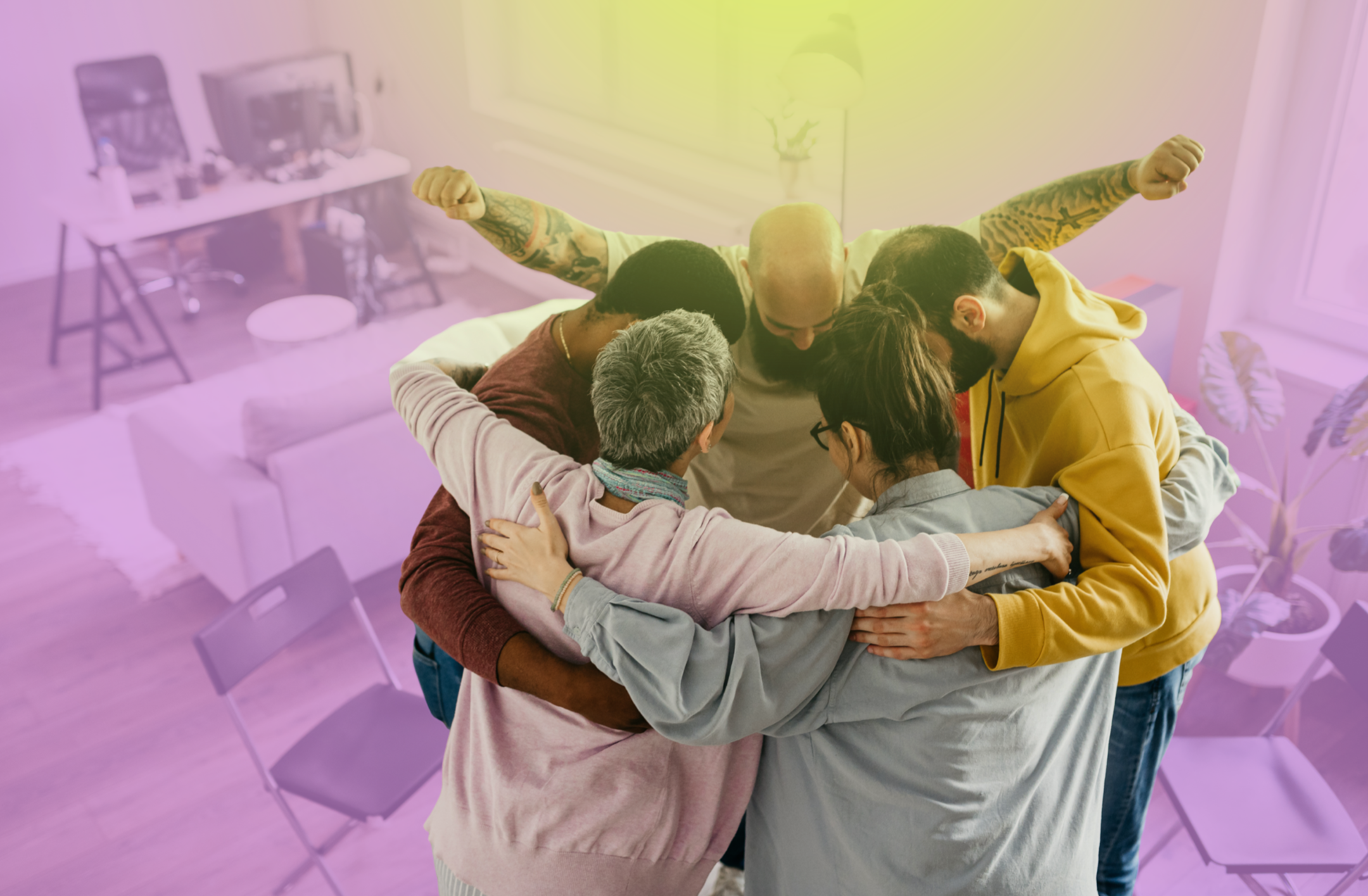 A diverse group of six people, including men and women of different ages and ethnicities, huddled together in a circle with arms around each other, displaying a moment of unity and support in a well-lit, cozy living room with modern furniture and houseplants.