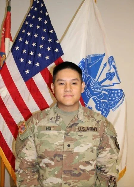 Young man in U.S. Army uniform standing in front of American and military flags.
