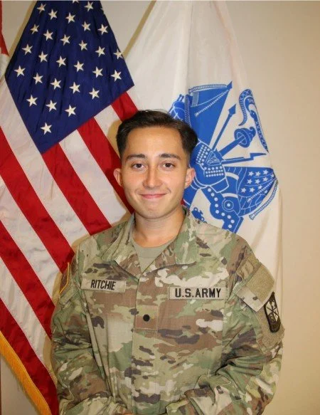 U.S. Army soldier in uniform standing in front of the American flag and a military flag, smiling.