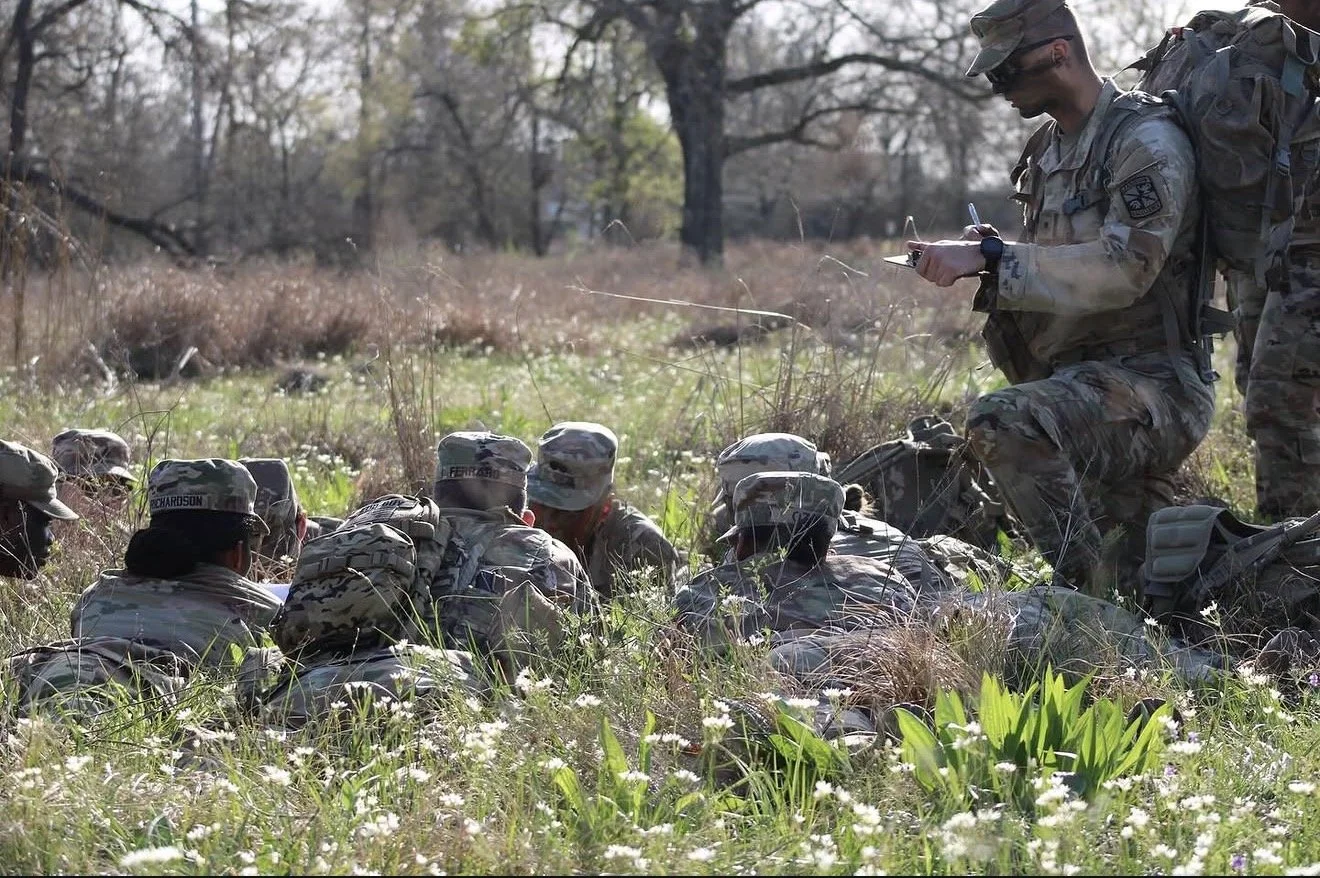 A group of soldiers in camouflage uniforms on the ground in a grassy field, with one soldier standing and writing in a notebook.