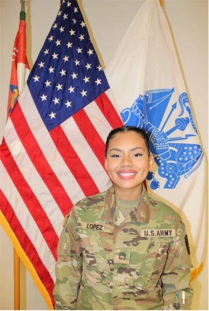 A female soldier in U.S. Army uniform standing in front of two flags, one American flag and one military flag.