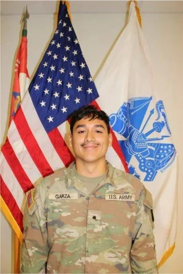 Young man in military uniform standing in front of American and military flags.