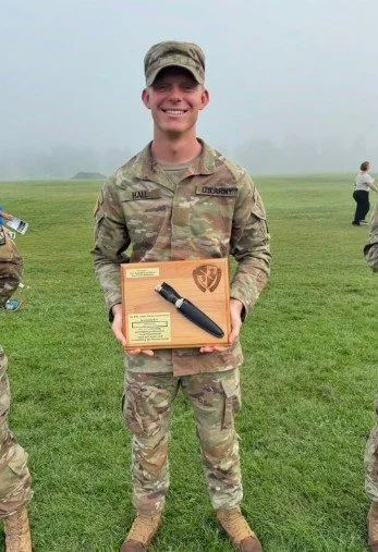 A smiling soldier in camouflage uniform holding a box with a knife in it, outdoors on a grassy field.