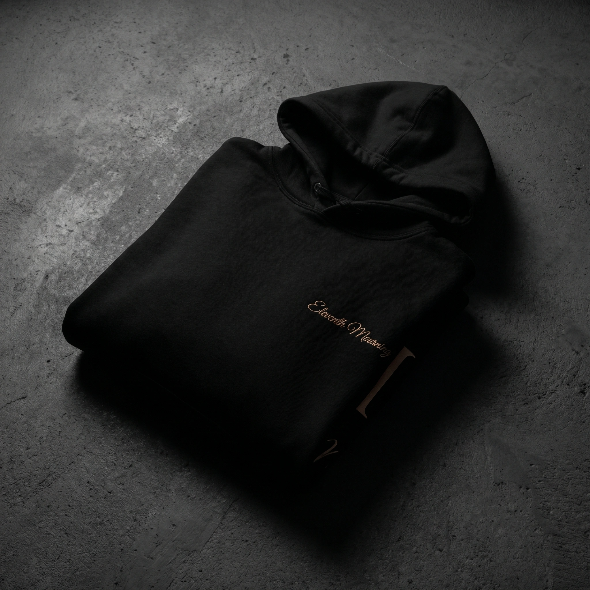 A black hoodie with partial embroidery on the front is placed on a gray textured concrete floor.