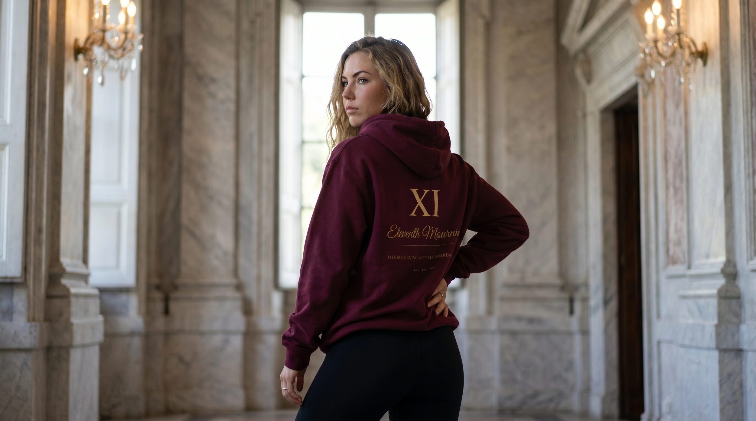 A woman with wavy light brown hair wearing a maroon hoodie with gold text on the back stands in an elegant, historic building with high ceilings, large windows, and marble columns.