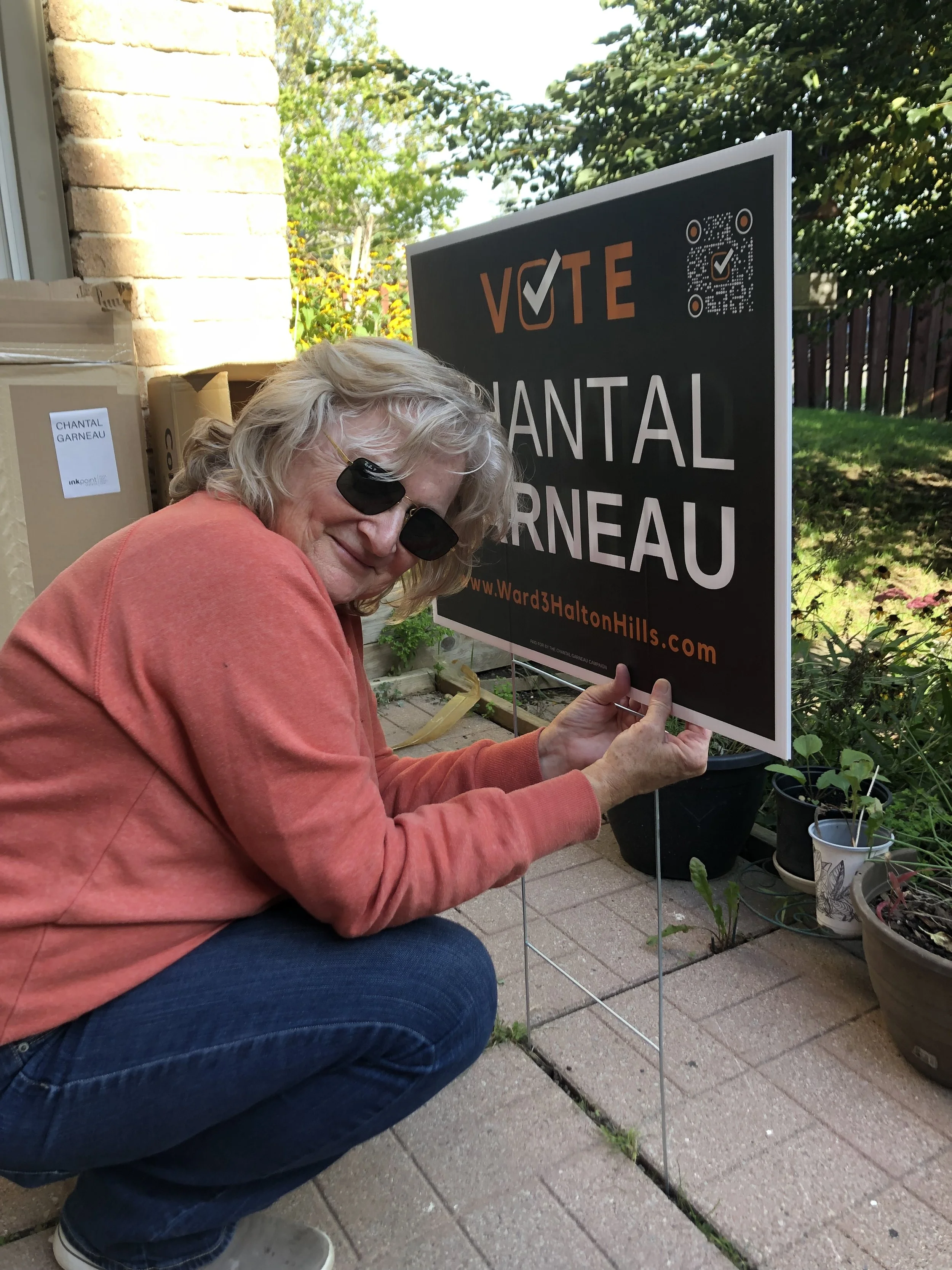 A woman with blonde hair wearing sunglasses, an orange sweater, and blue jeans squats on the patio holding a protest sign that reads 'VOTE CHANTAL ARNEAU' with a website link at the bottom. She is smiling and in front of plants and boxes.