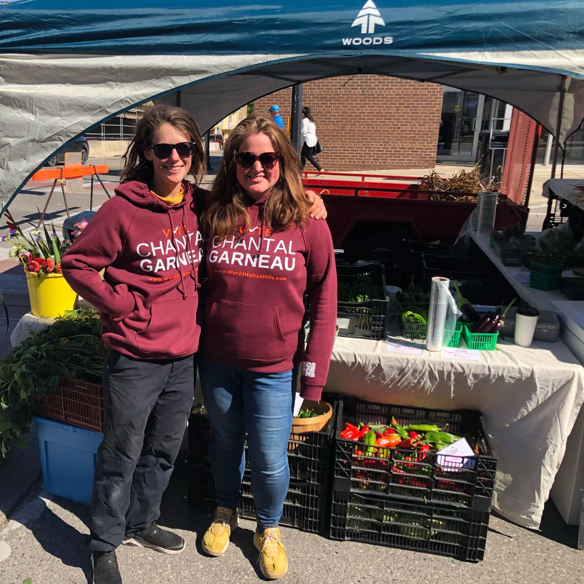 Two women standing in front of a market stall with vegetables, wearing maroon hoodies and sunglasses, outside on a sunny day.
