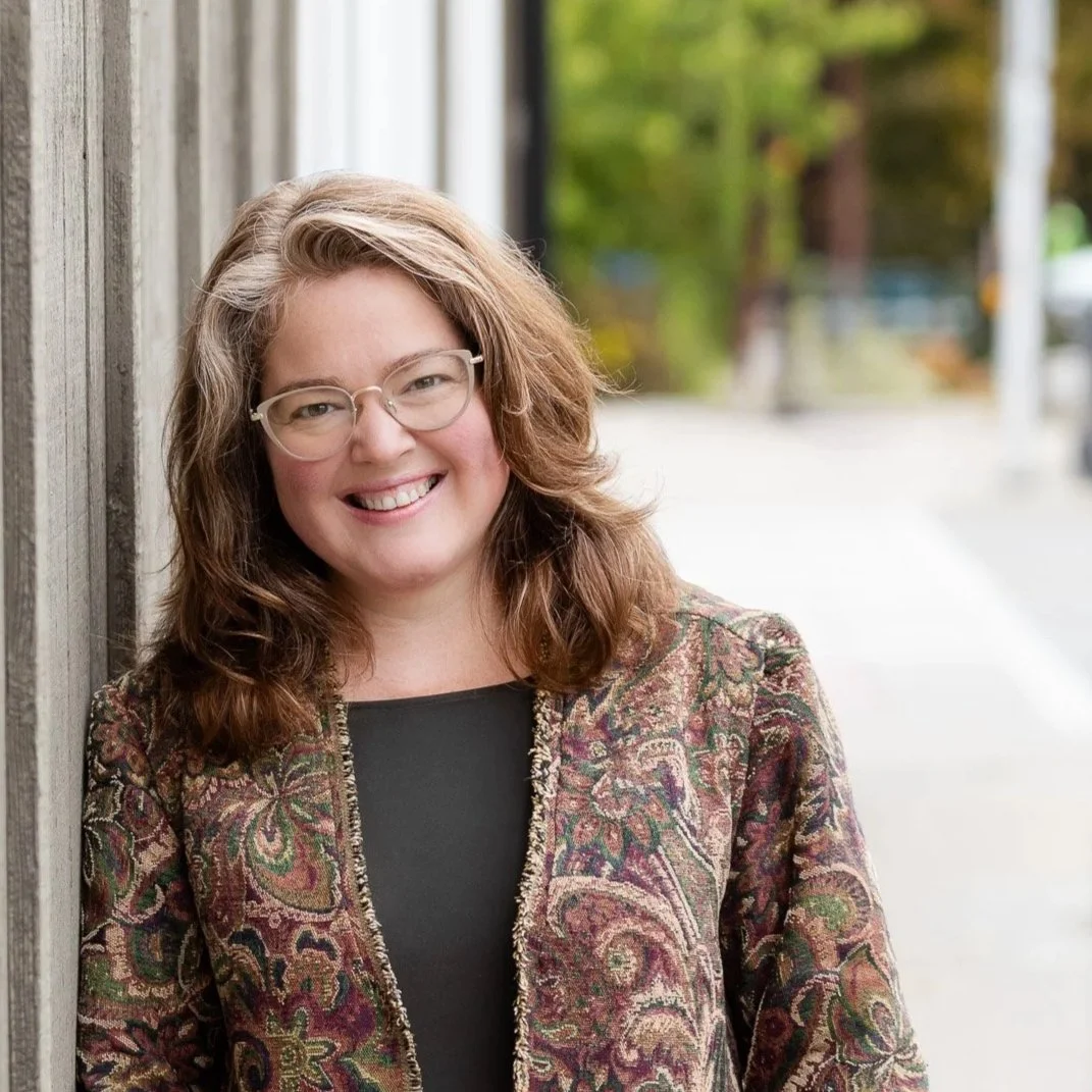 A woman smiling and wearing glasses, standing outside leaning against a wooden wall, with blurred trees and sidewalk in the background.