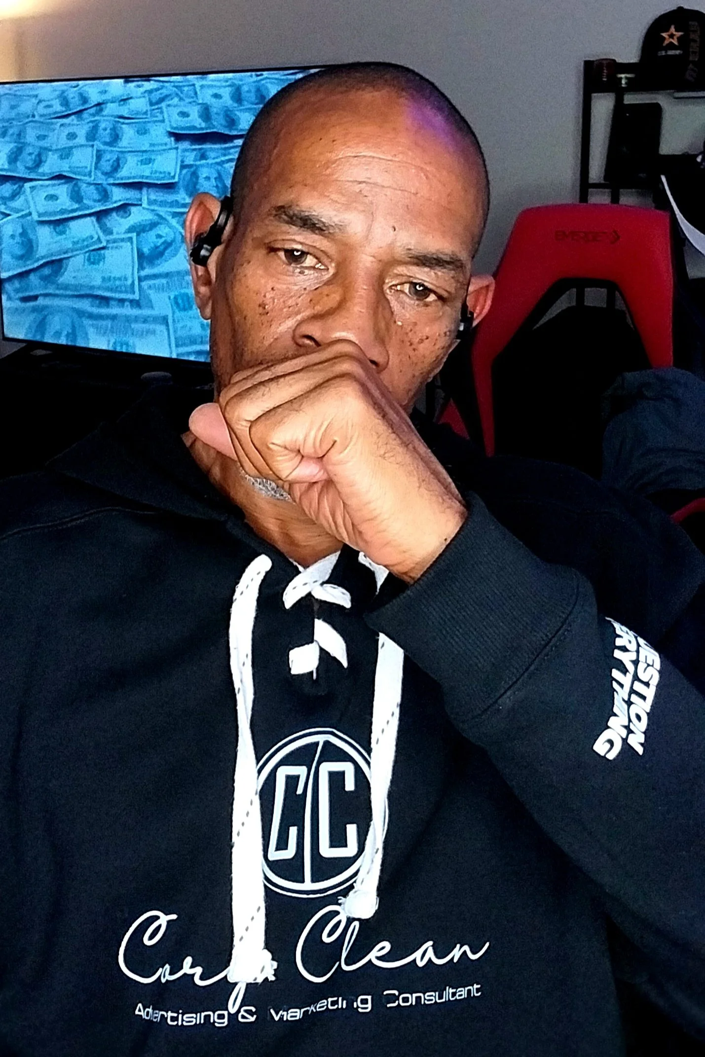 Cory Clean with a shaved head and freckles, wearing a black hoodie with white text, sitting indoors with a hand covering his mouth, in front of a television screen displaying blue-tinted US dollar bills.