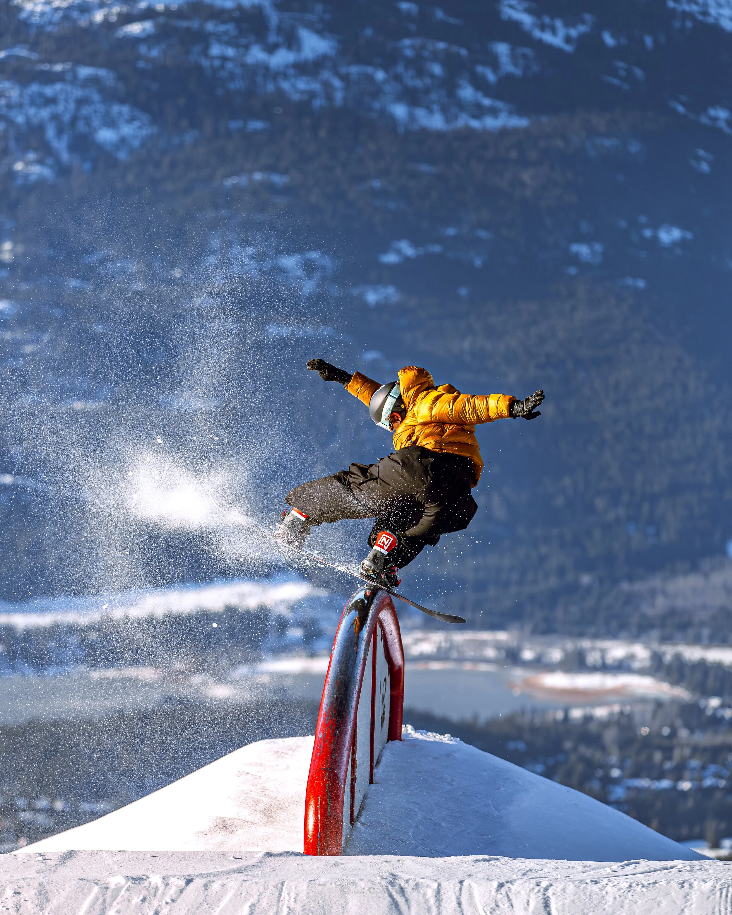 A person in a yellow jacket and gray ski pants snowboarding on a snow-covered mountain, with mountains in the background and clear blue sky.