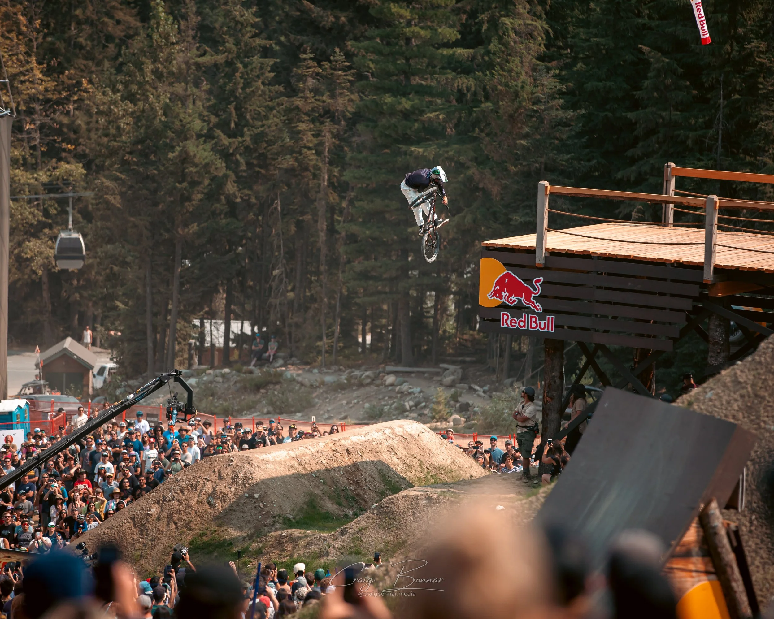 A mountain biker performing a jump off a wooden ramp with a Red Bull logo, while a crowd of spectators watches in a forested outdoor event area.