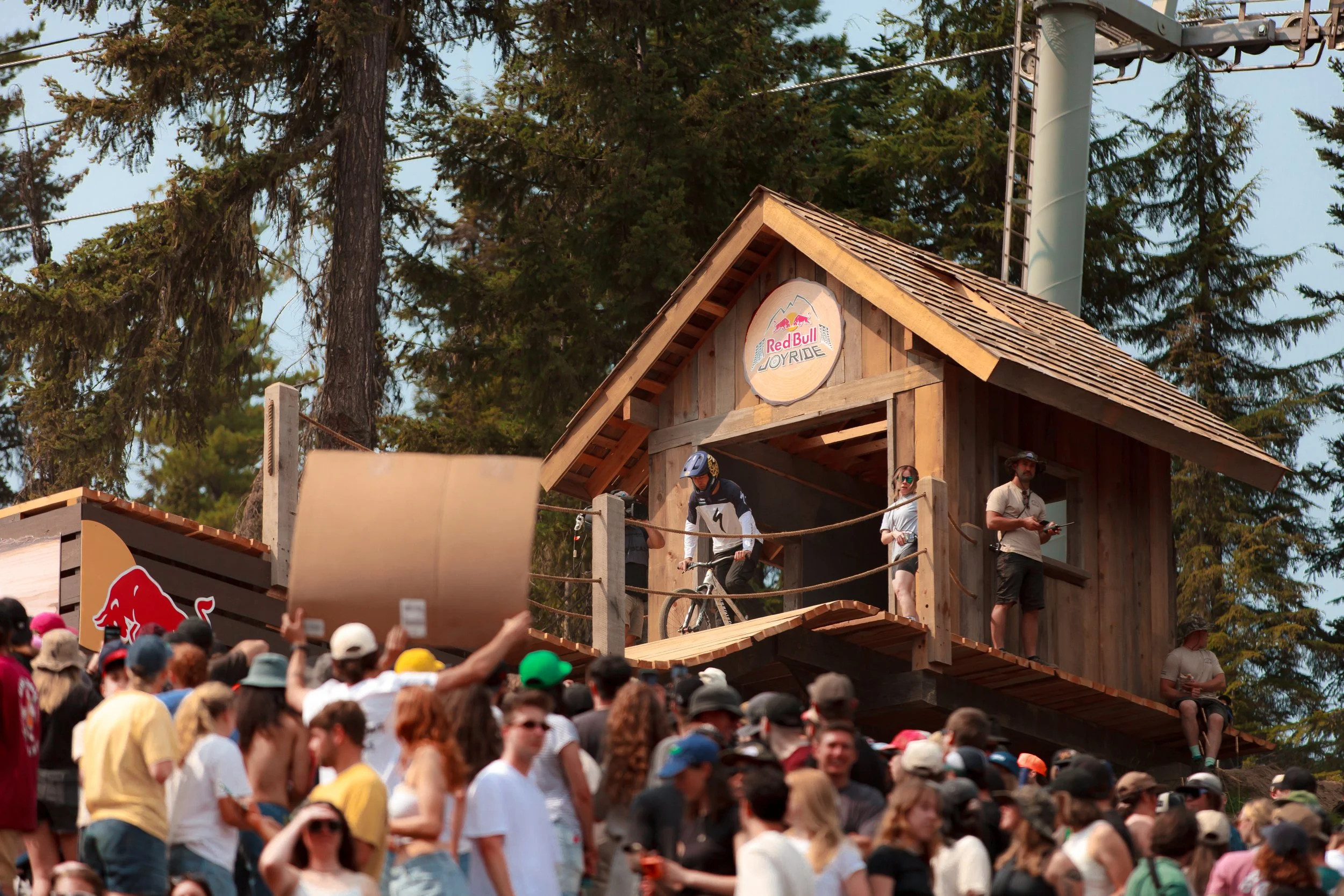 Crowd of people gathered at an outdoor event with a wooden structure labeled 'Red Bull Joyride' and people preparing to ride bicycles on a ramp, surrounded by tall pine trees.