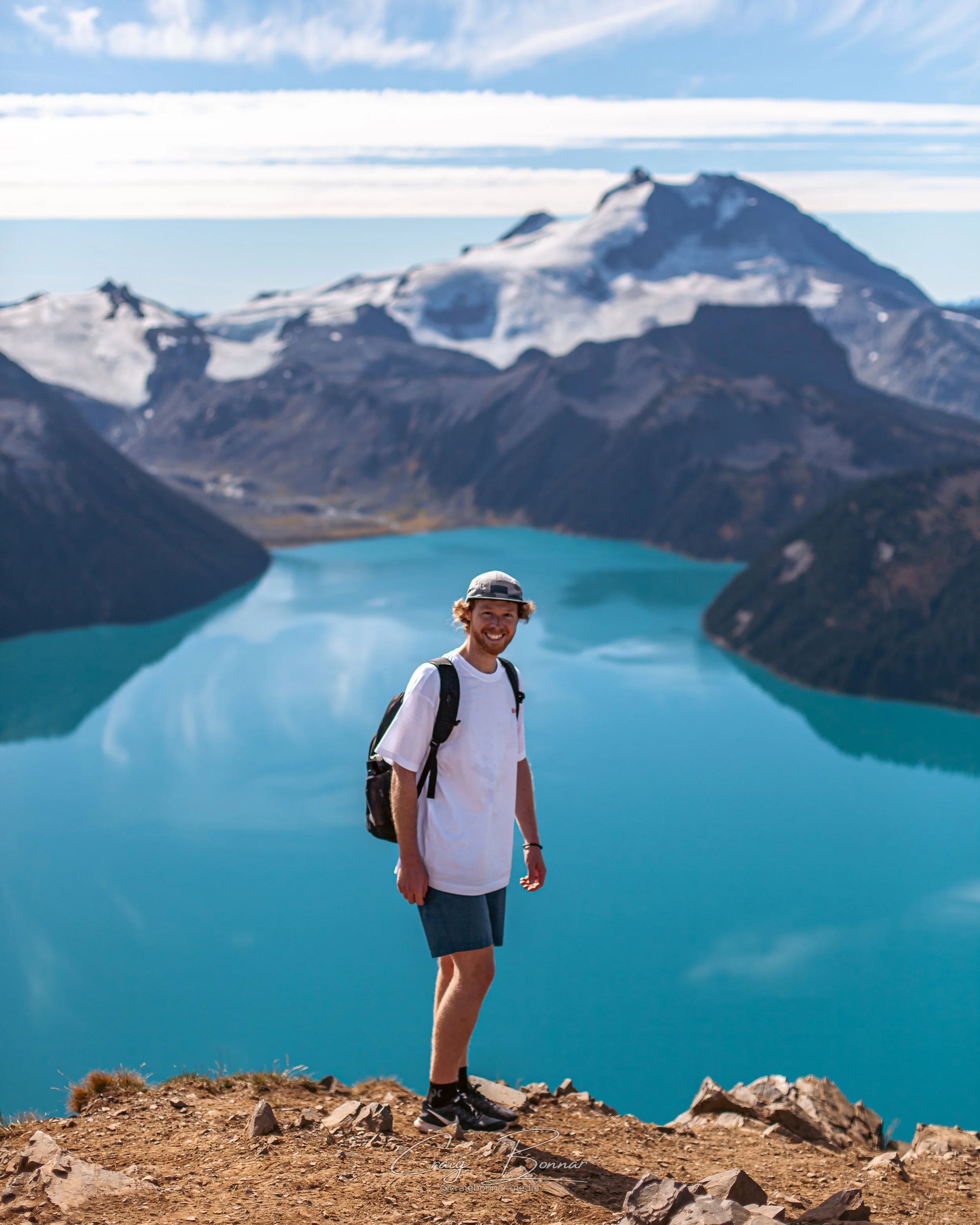 A man standing on rocky ground by a blue lake with snow-capped mountain in the background.