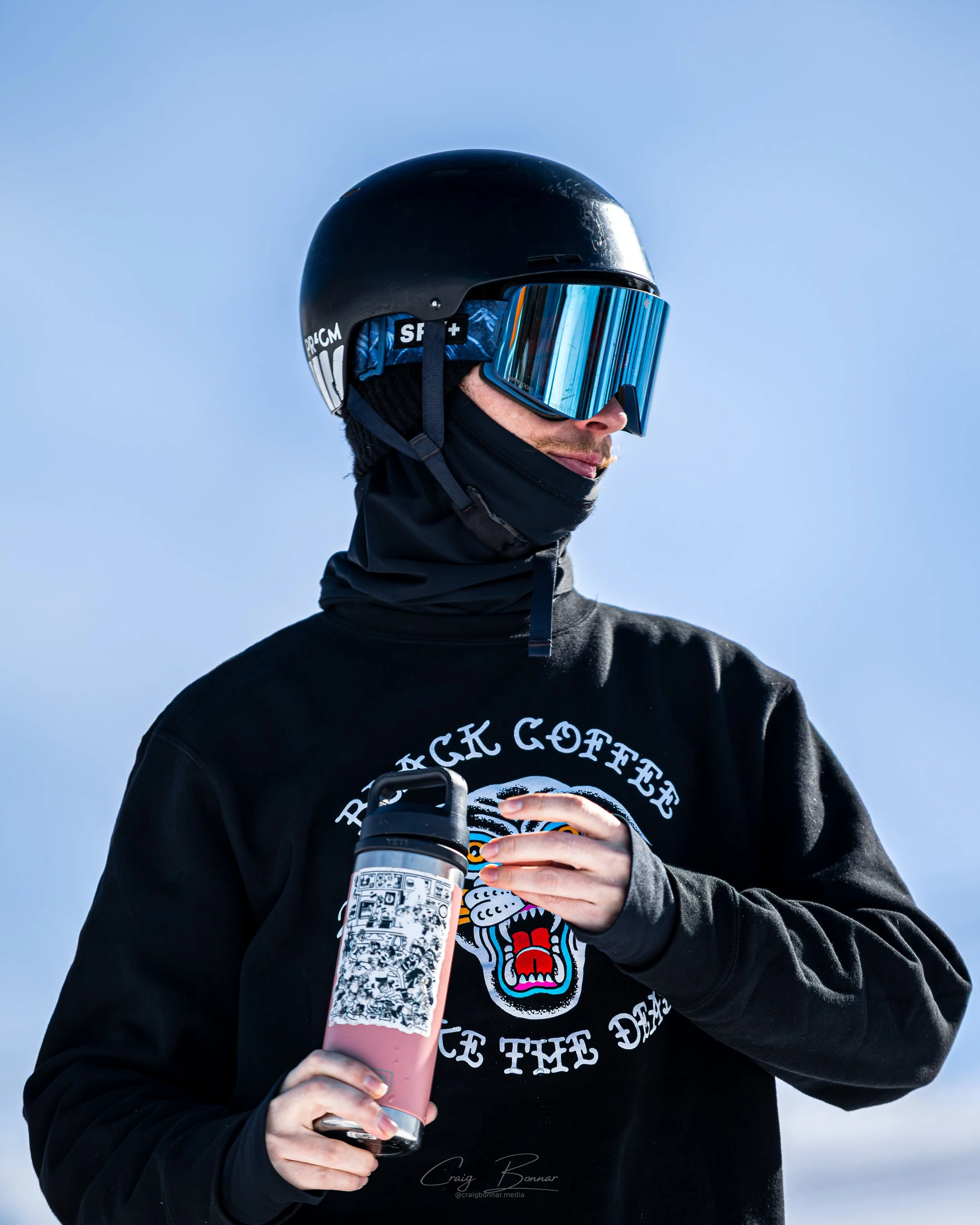 Person wearing a black helmet with reflective goggles, black jacket, and graphic shirt, holding a pink insulated water bottle with black cap against a clear blue sky.