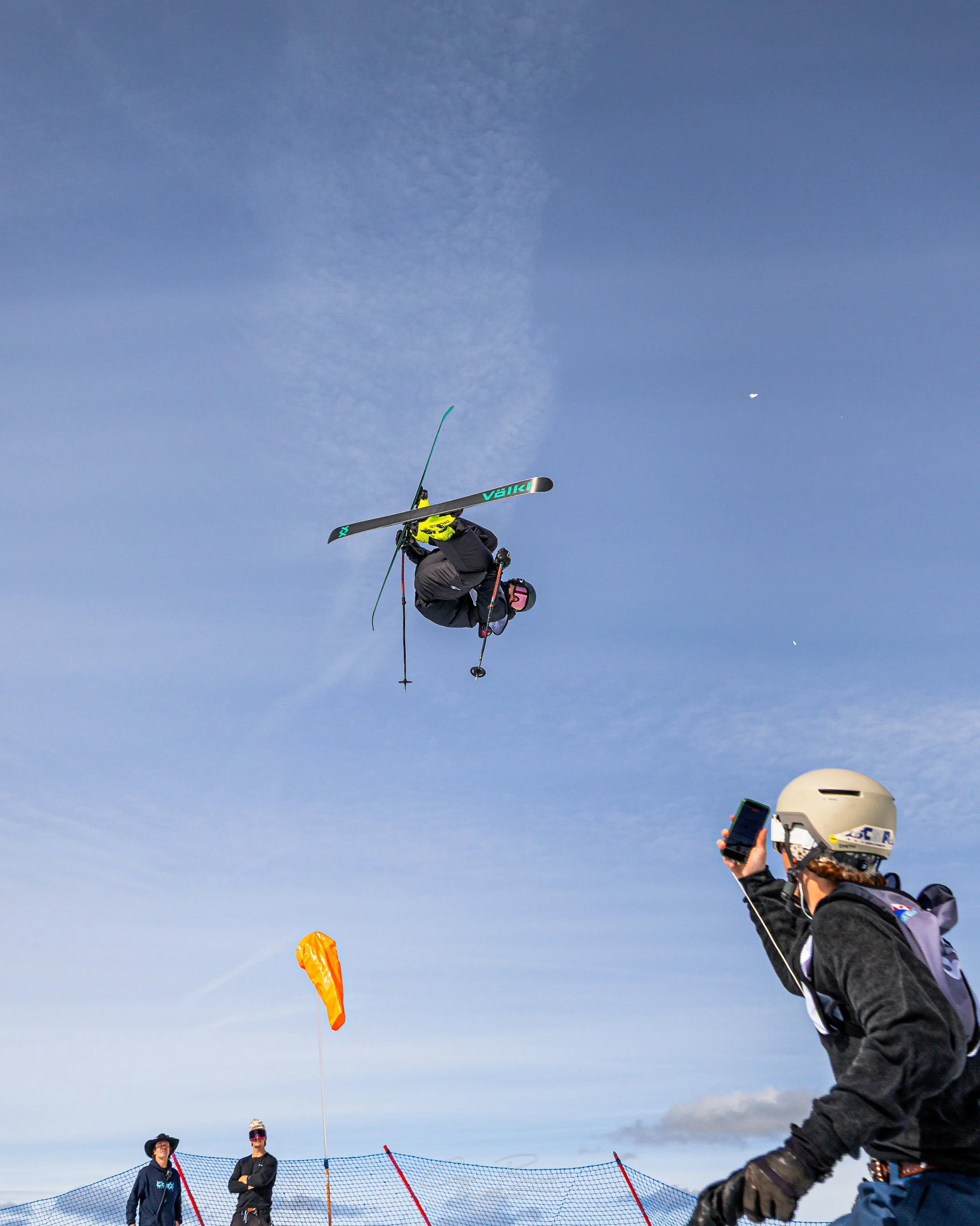 People on a snowy mountain participating in a winter sport, with some standing near a safety net and others in mid-air performing tricks on skis, under a bright blue sky.
