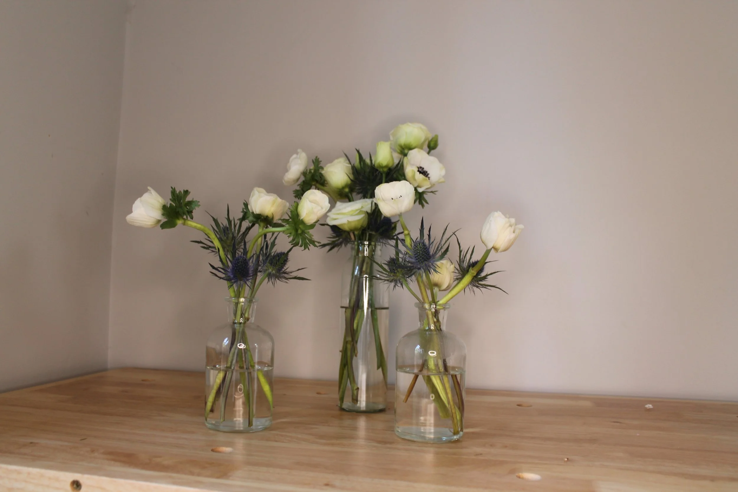 Three small glass vases with white and purple flowers on a wooden surface against a plain wall.