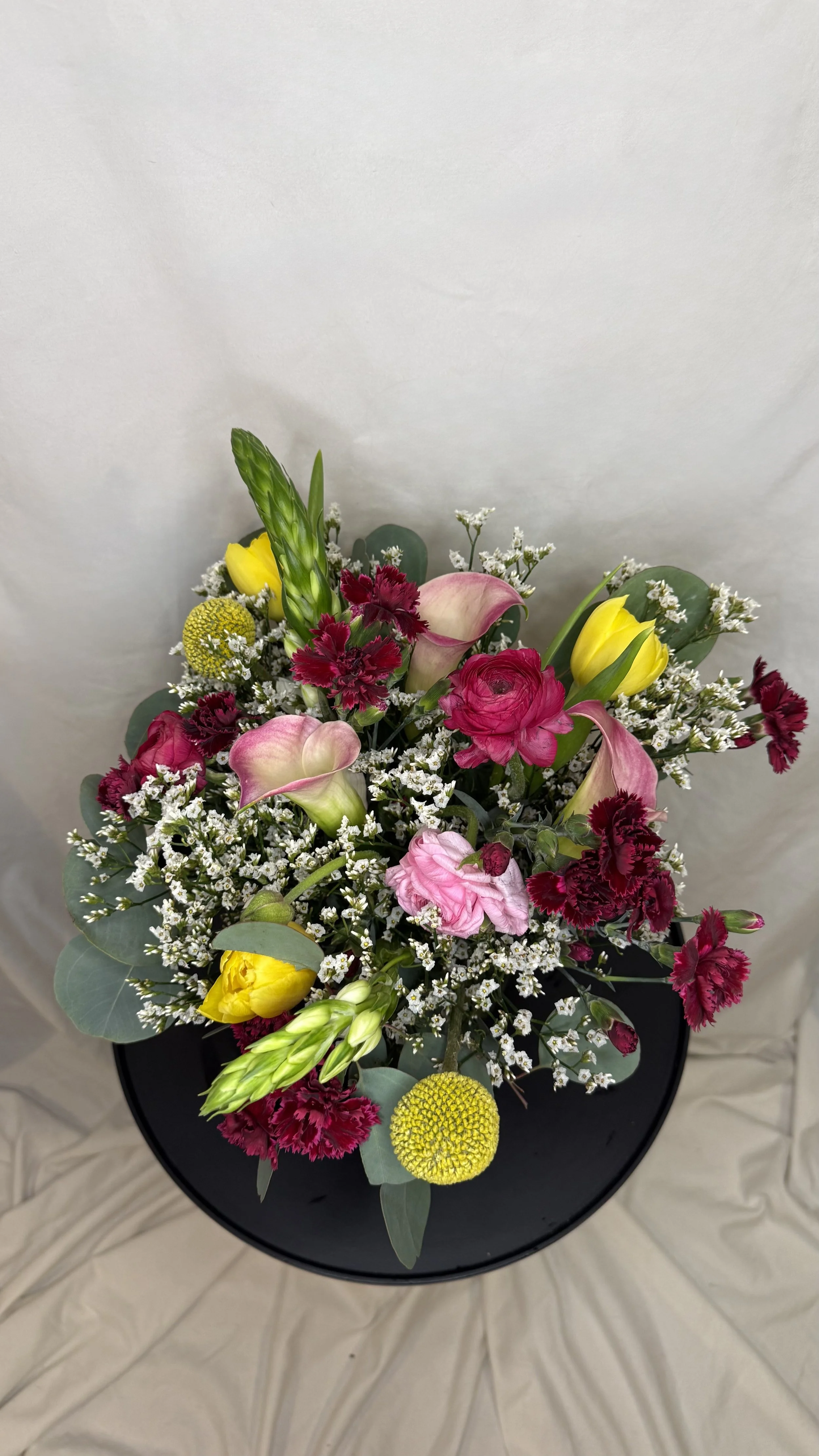 A colorful bouquet of flowers in a black pot, featuring pink calla lilies, red carnations, yellow tulips, white baby's breath, green eucalyptus leaves, and yellow craspedia, arranged on a white fabric surface.