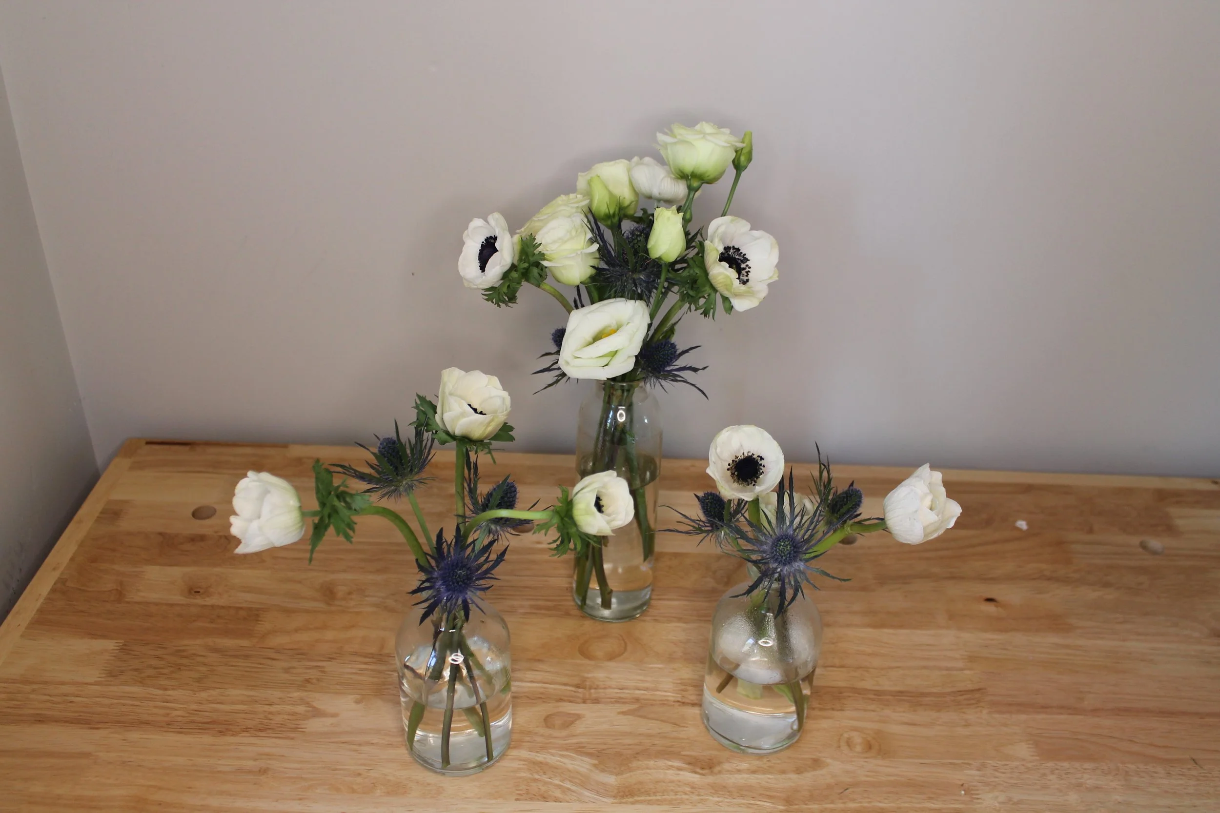 Four clear vases with white and purple flowers sit on a wooden table against a plain light-colored wall.