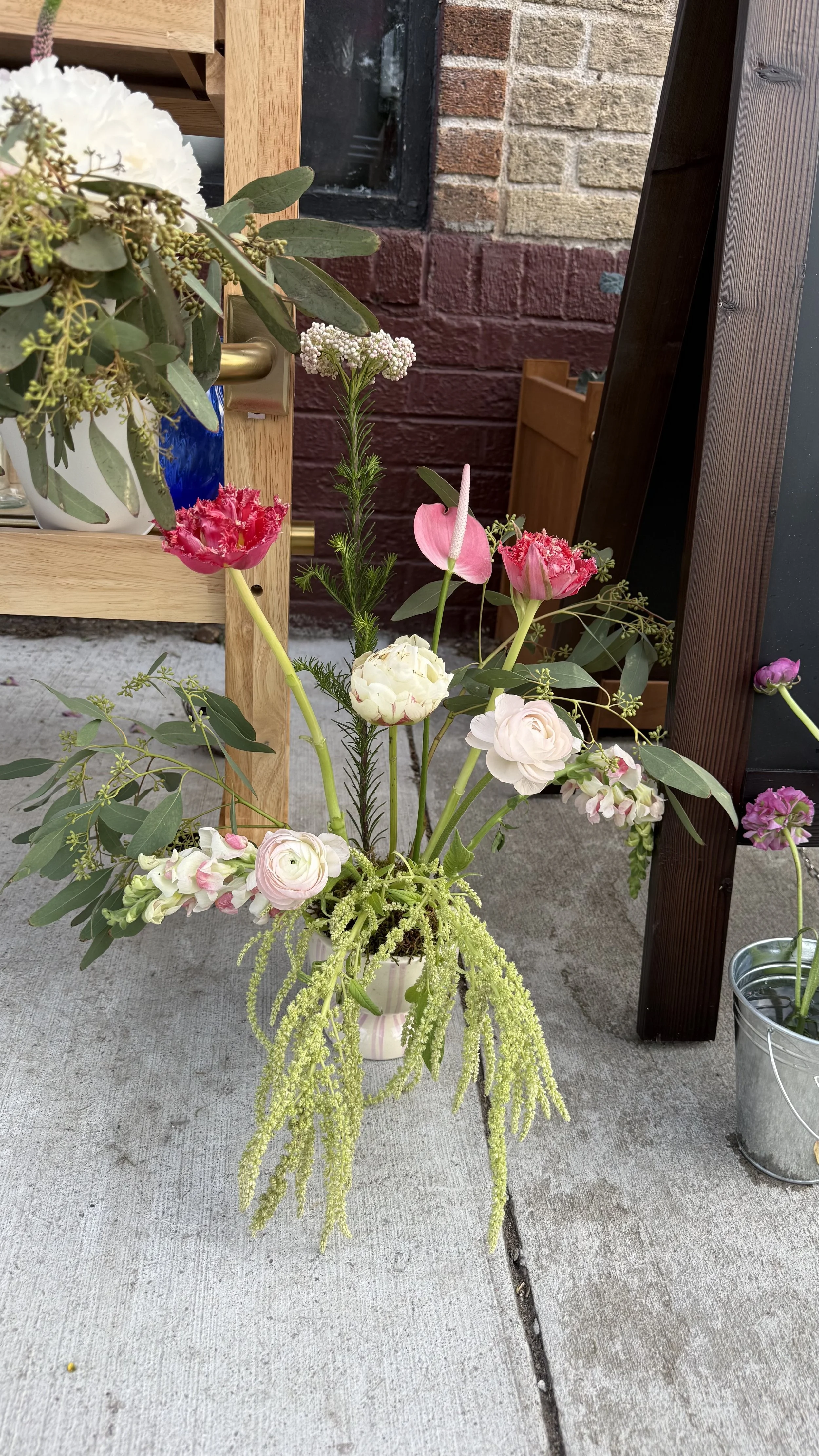 A vibrant floral arrangement in a white vase containing pink, white, and cream-colored flowers with green foliage, placed on a concrete surface outdoors near a brick wall.