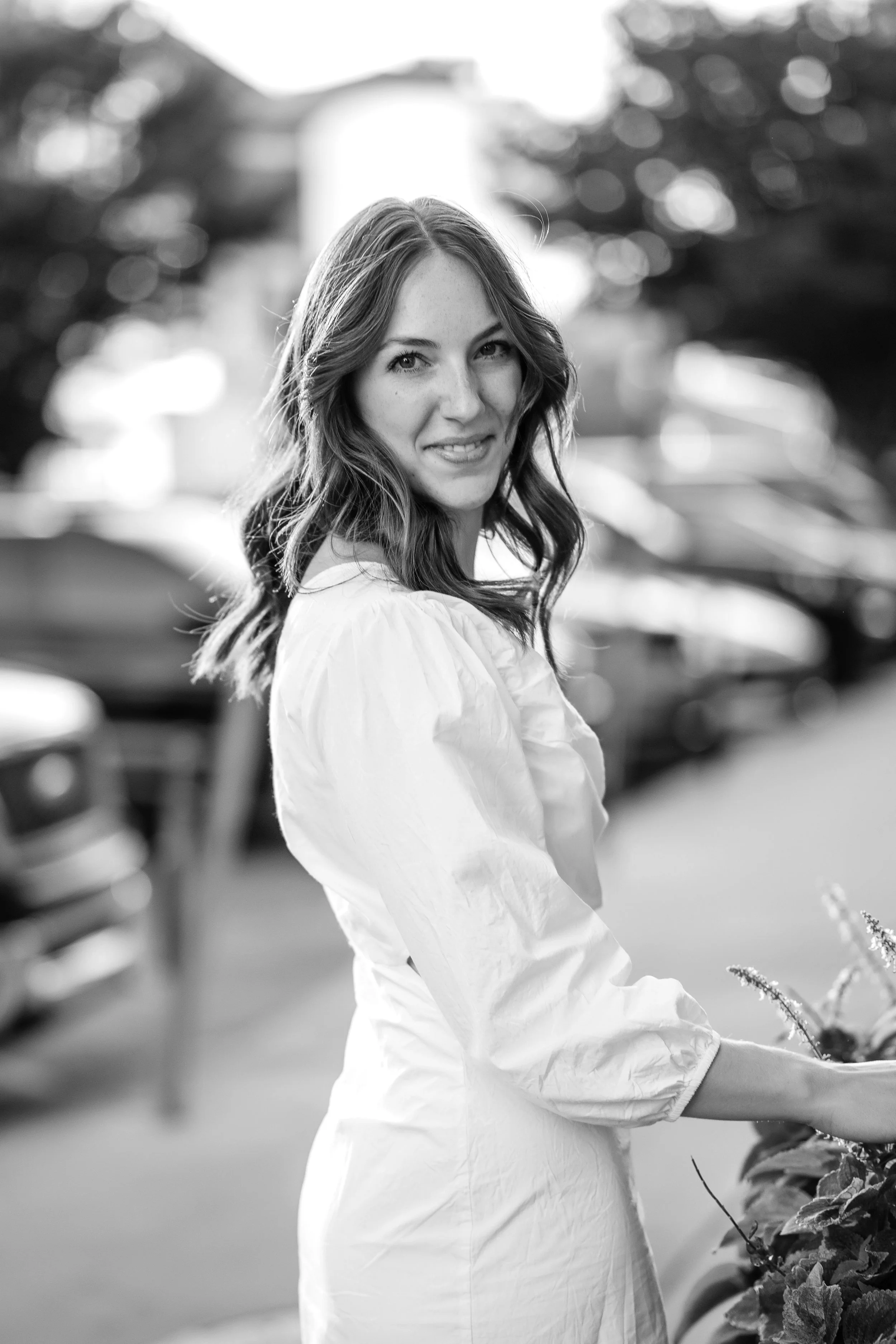 A woman with wavy hair smiling, wearing a light-colored blouse, standing outdoors with blurred cars and trees in the background.