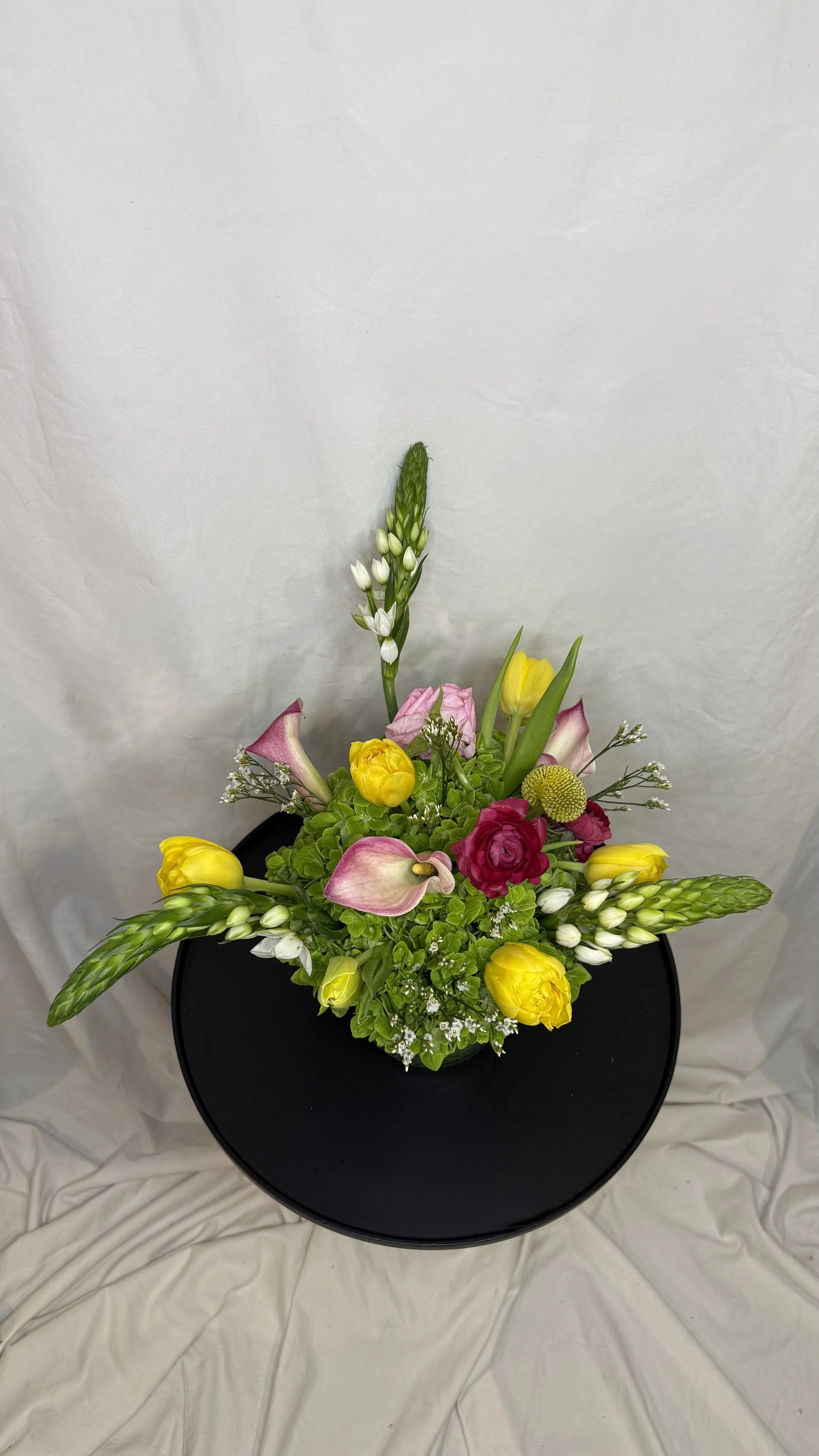 A flower arrangement in a black round vase with yellow, pink, white, and green flowers, including calla lilies, tulips, and greenery, arranged on a black round table against a plain white background.