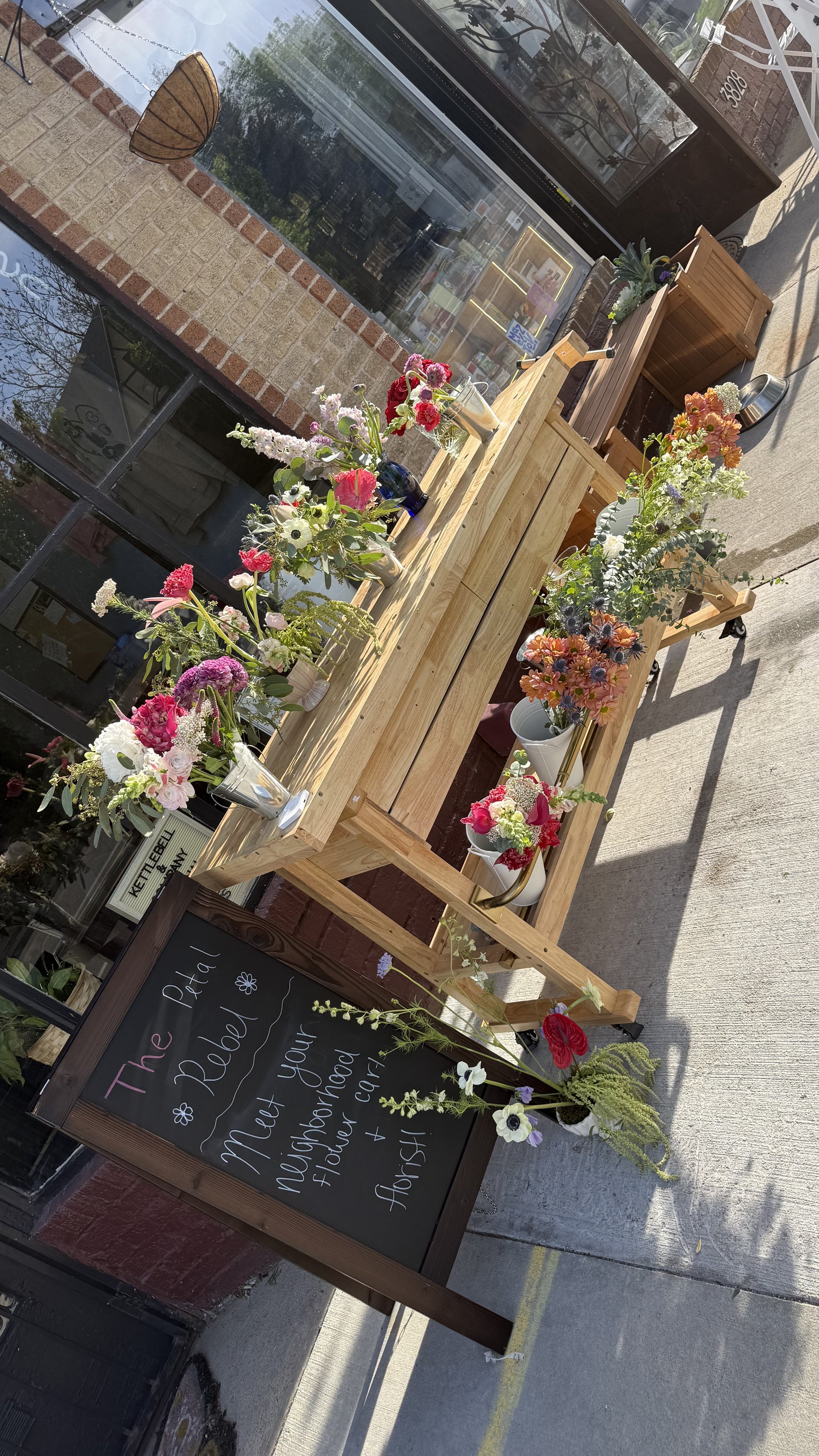 Wooden flower display stand with various colorful flowers outside a store, next to a chalkboard sign with handwritten message.