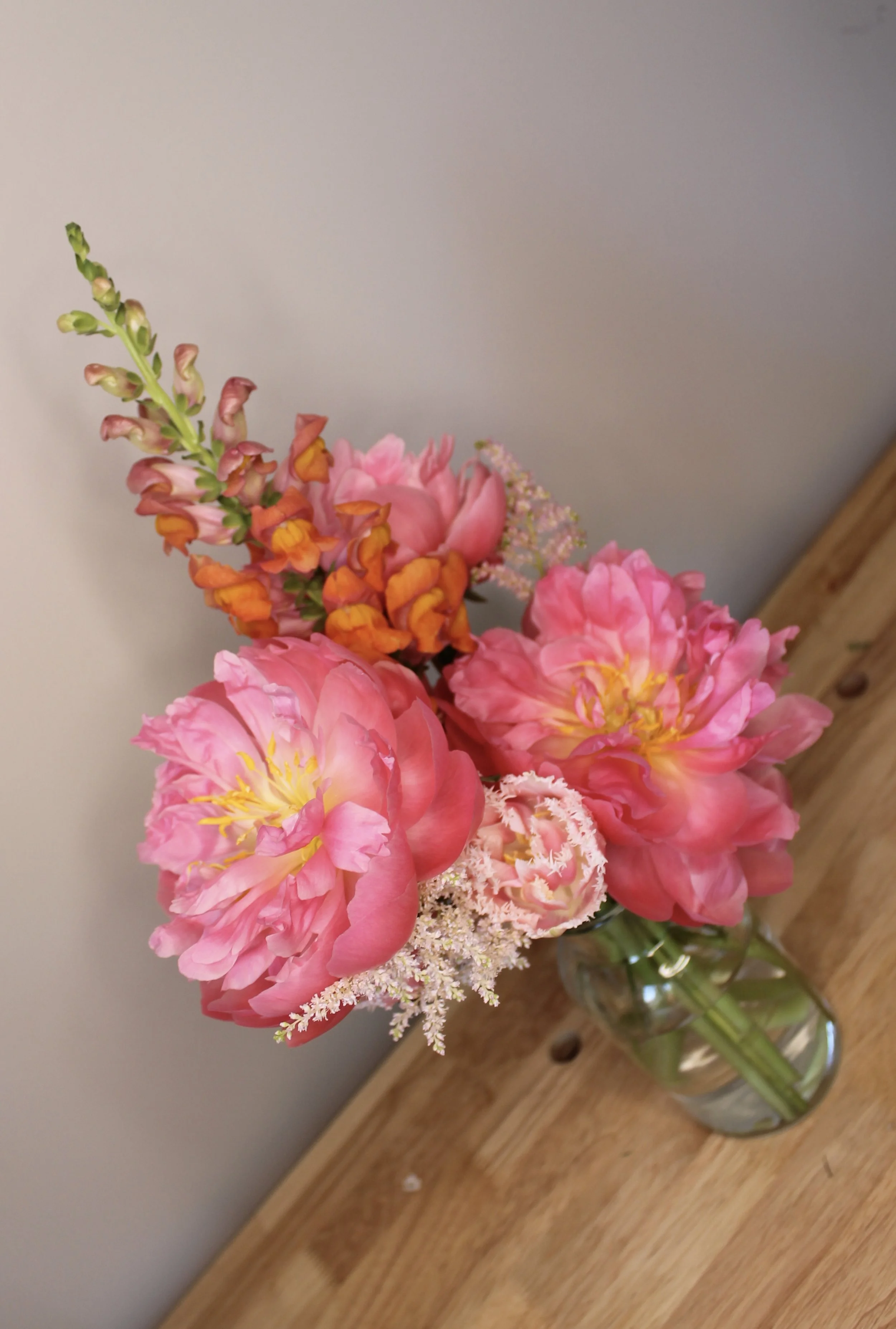 A bouquet of pink peonies and orange flowers in a glass jar on a wooden surface.