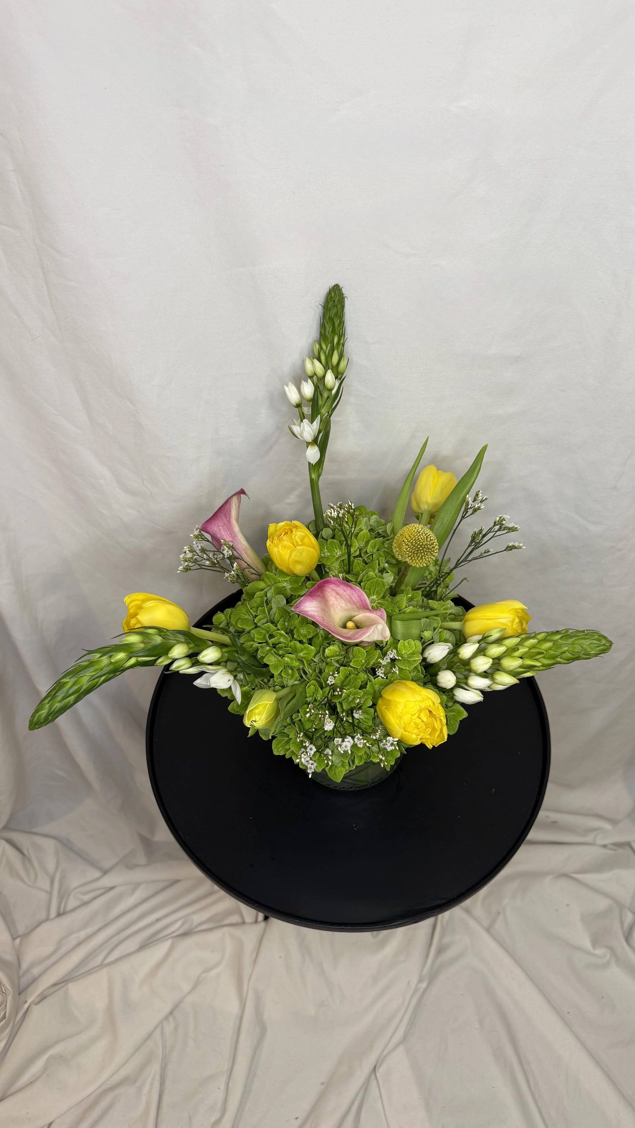 A floral arrangement with yellow tulips, pink calla lilies, white snapdragons, small white filler flowers, and green foliage in a black pot on a black round table against a white backdrop.