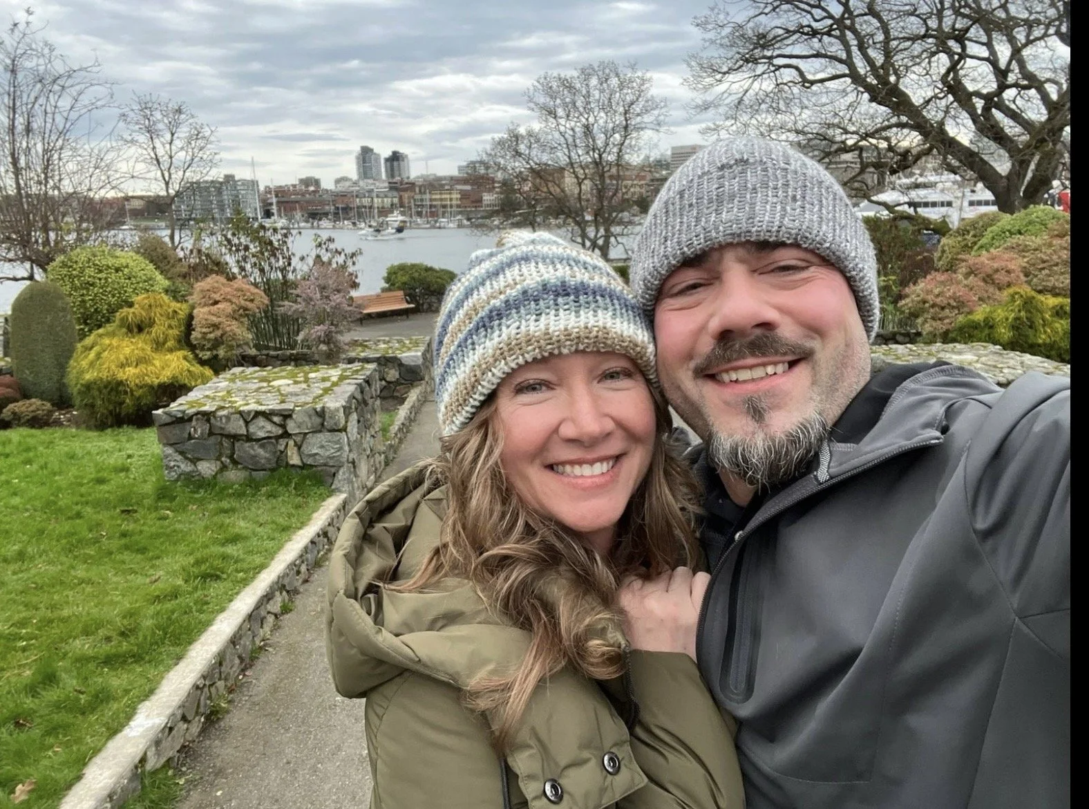 A smiling couple in warm clothing taking a selfie outdoors near a garden with trees and shrubs, with a river, boats, and city buildings in the background on a cloudy day.