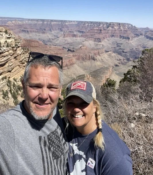A smiling couple taking a selfie at the Grand Canyon with the canyon in the background on a sunny day.