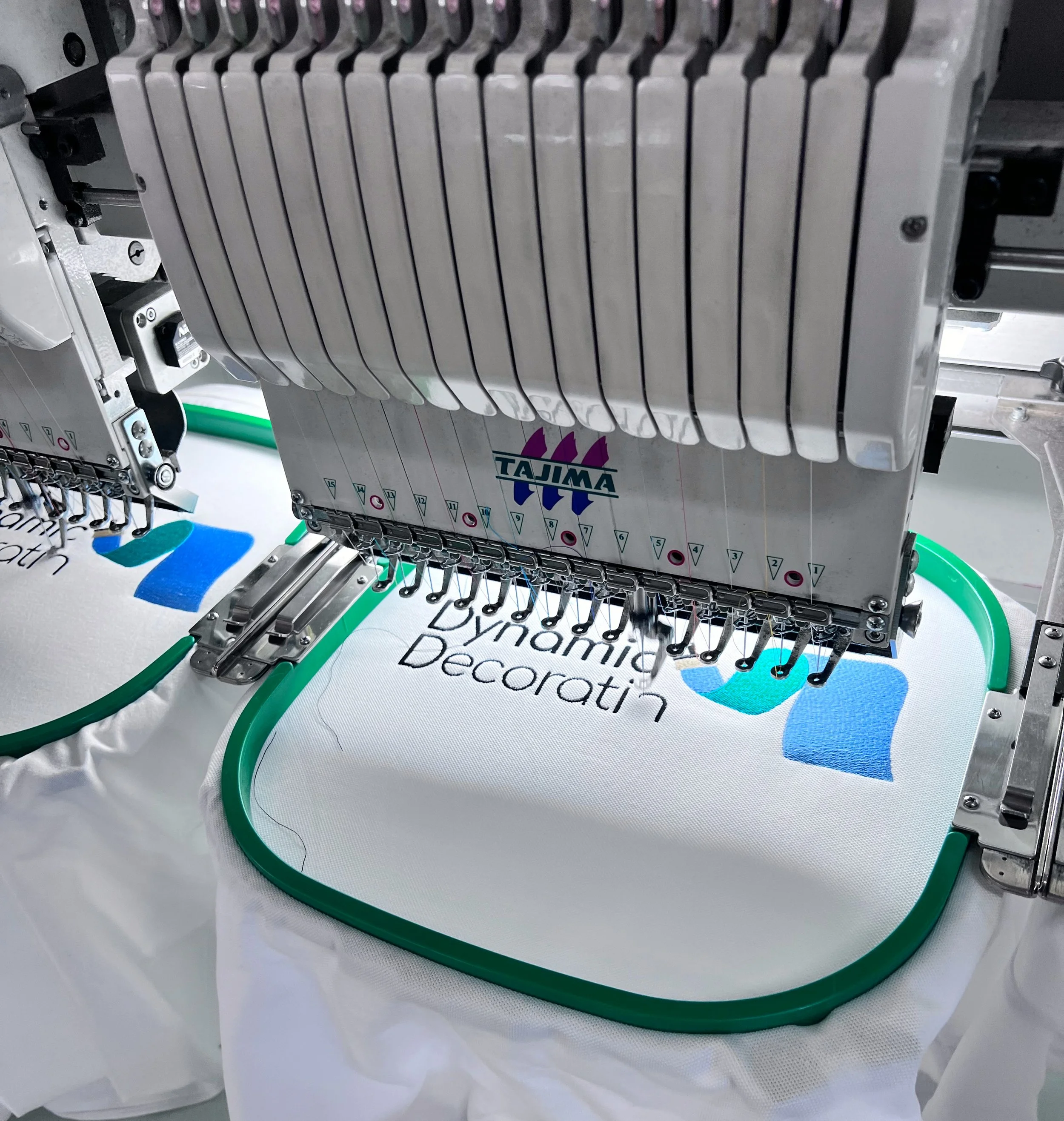 A close-up of an embroidery machine stitching a design on fabric with a green hoop, displaying part of the text 'Dynamic Decorati' and colorful shapes.
