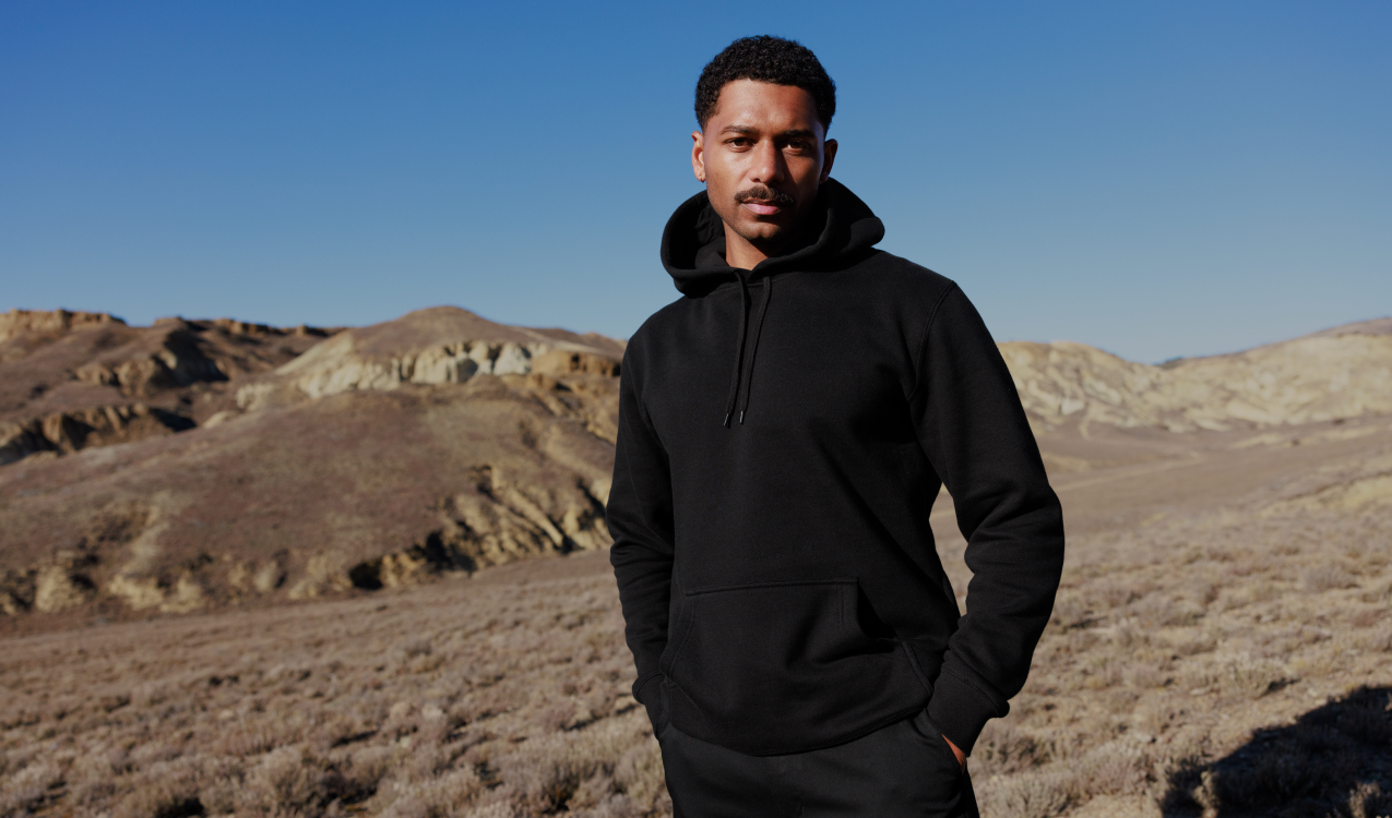 A young man with short curly hair and a thin mustache stands outdoors in a desert landscape with hills in the background, wearing a black hoodie and looking at the camera.