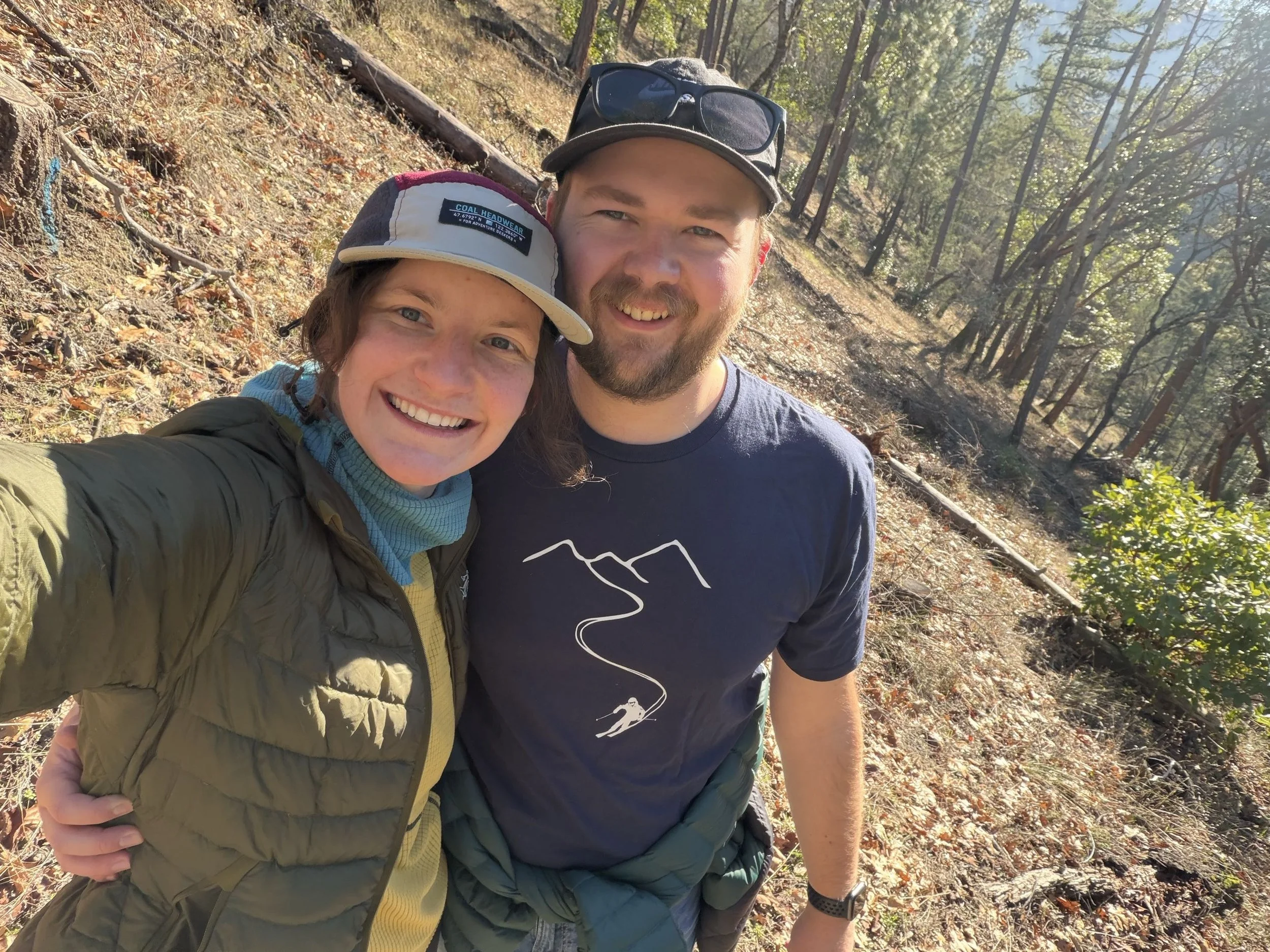 A smiling man and woman taking a selfie in a wooded outdoor area on a sunny day, wearing casual and outdoor clothing.