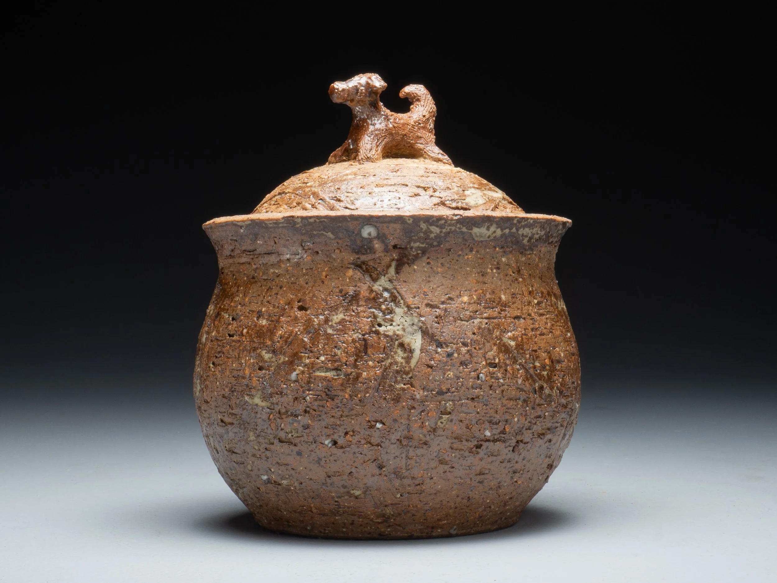 Ancient brown ceramic vessel with a small animal figure on the lid, set against a black background.