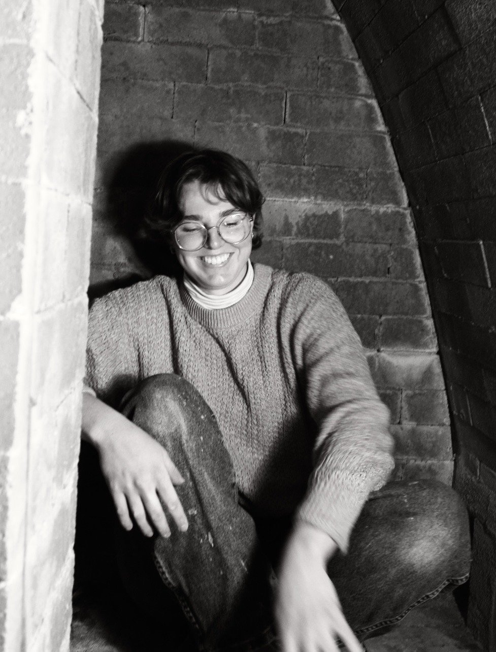 A smiling woman with glasses and short hair sitting in a corner between brick walls, wearing a sweater and jeans.