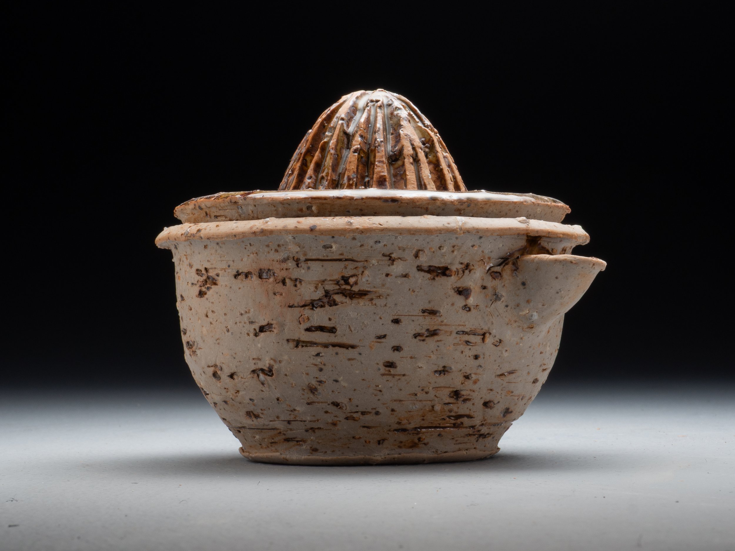 Close-up of a textured ceramic bowl with a lid, featuring a small, dome-shaped knob, on a neutral surface with a black background.