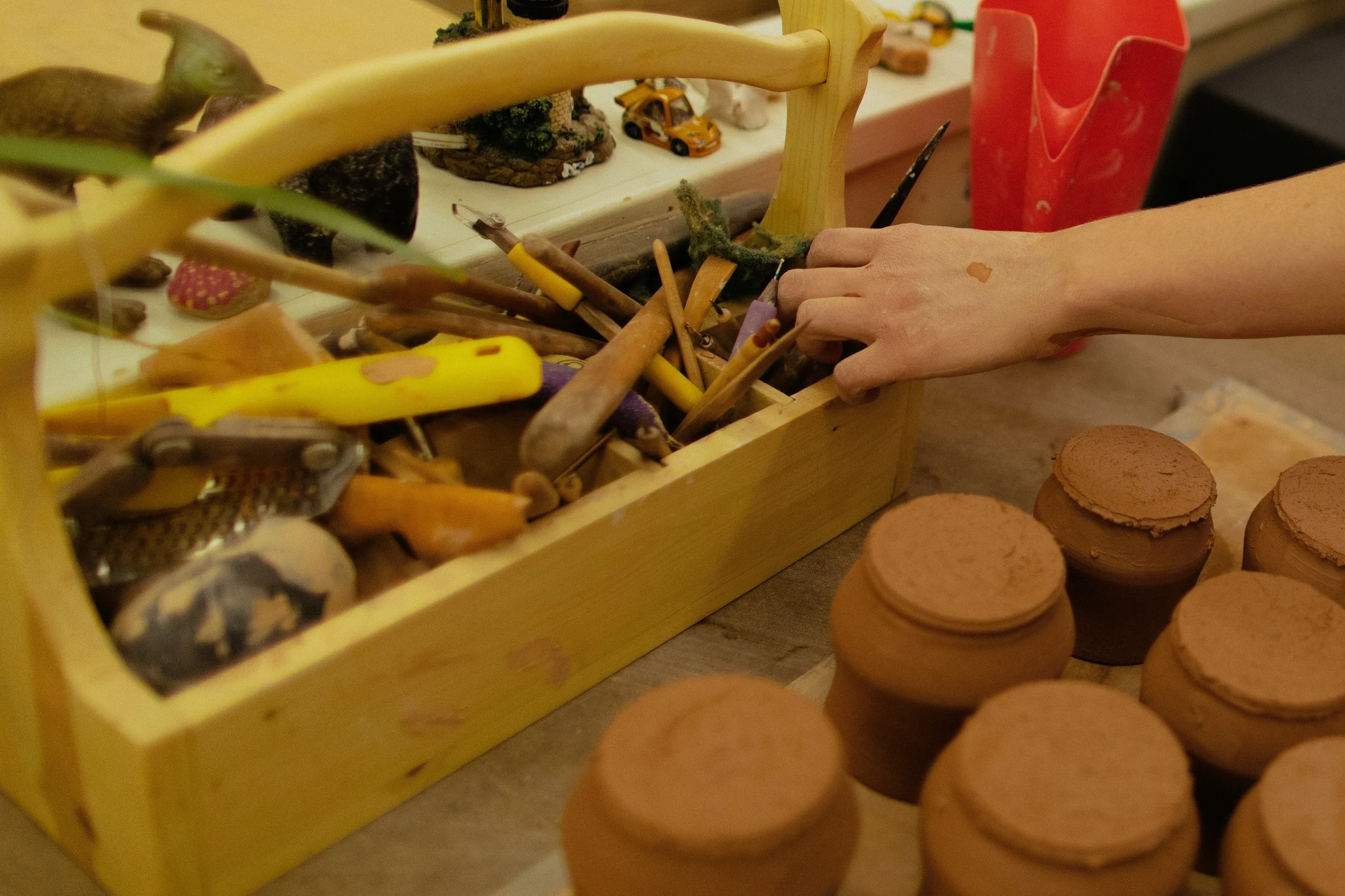 Wooden box filled with various miniature toys and figurines, surrounded by clay discs and a person's hand nearby.