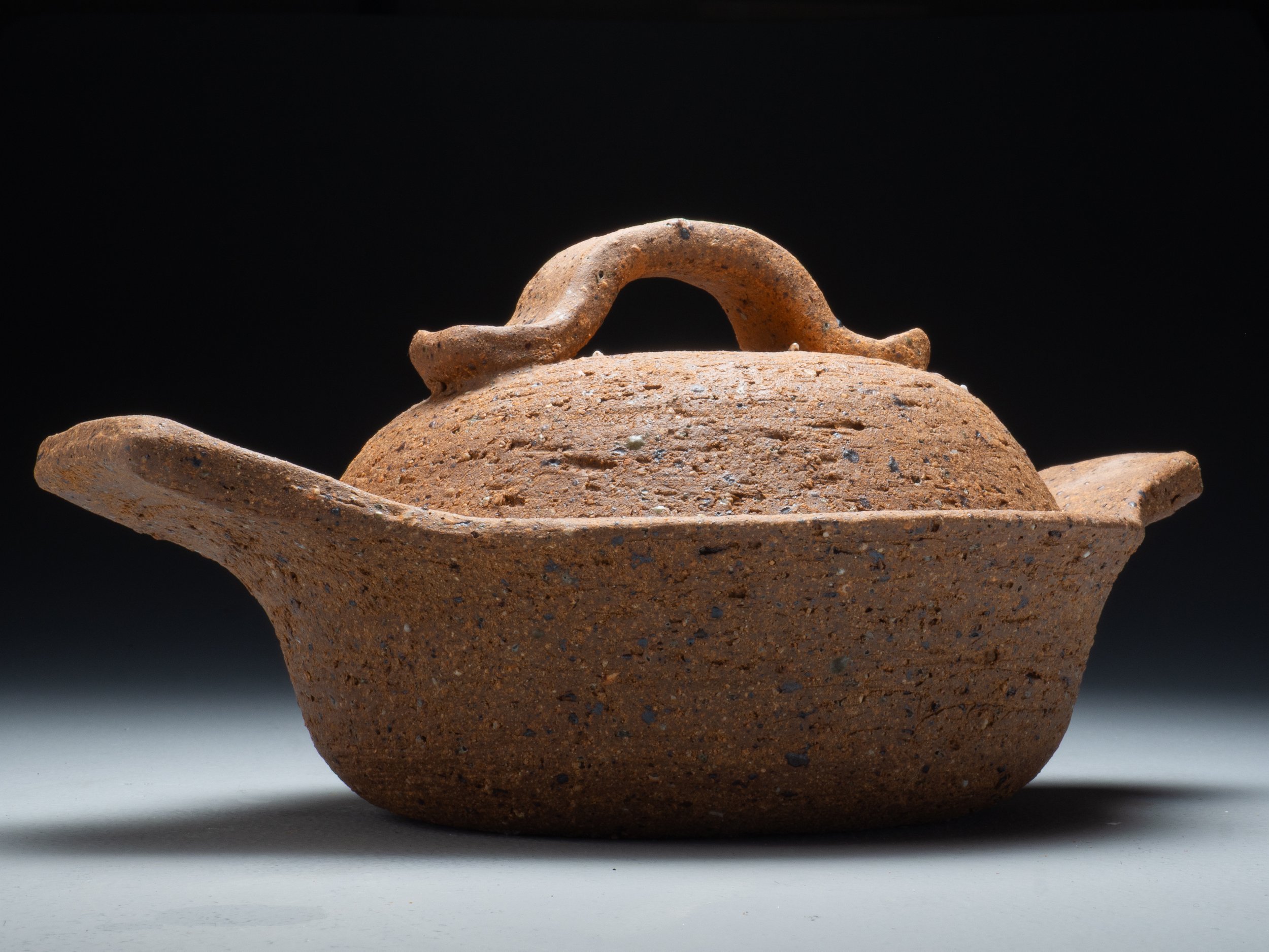A close-up image of an ancient clay oil lamp with a curved handle, placed against a dark background.