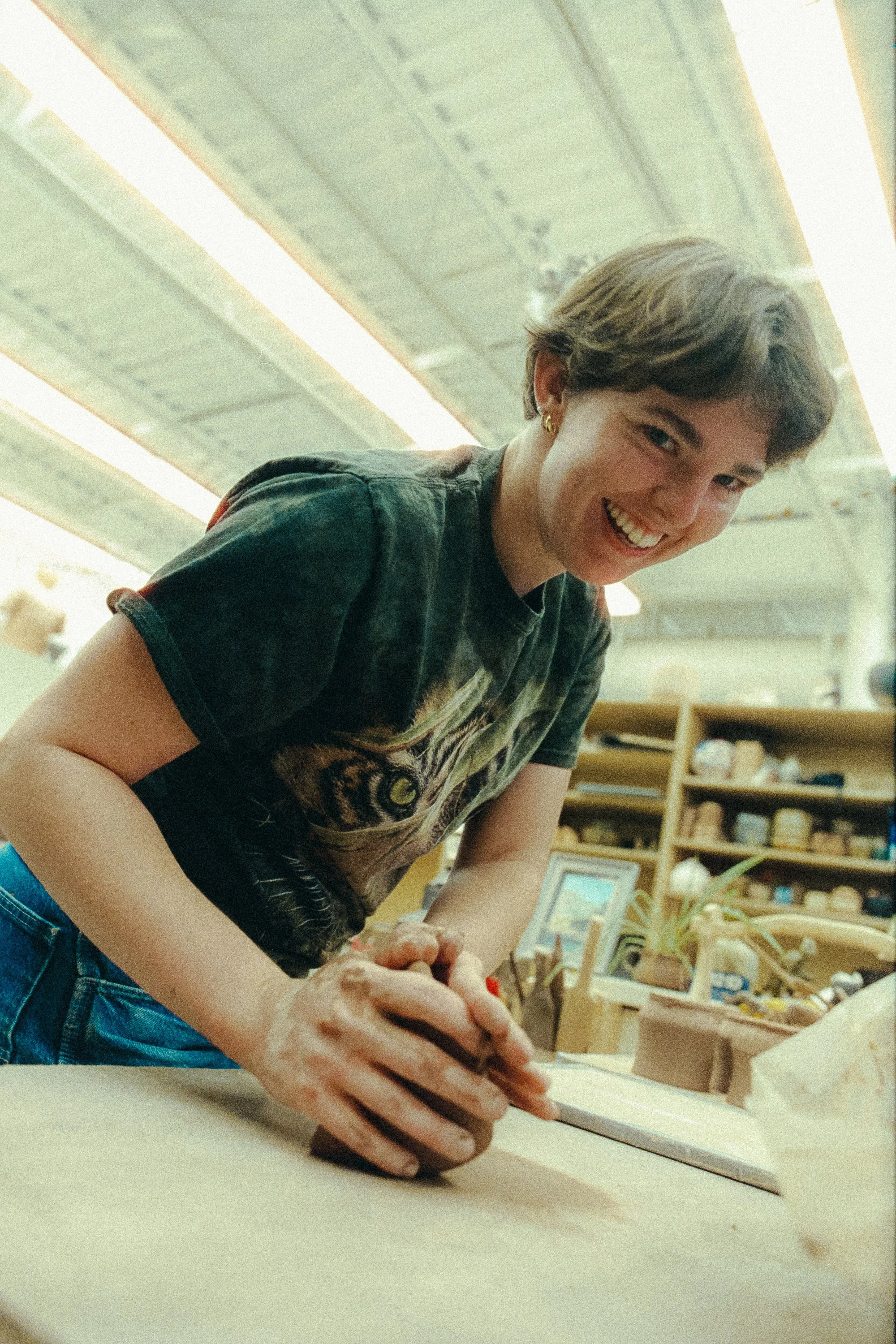 Young woman smiling while working on a craft project at a table in a workshop or classroom, with shelves and art supplies in the background.