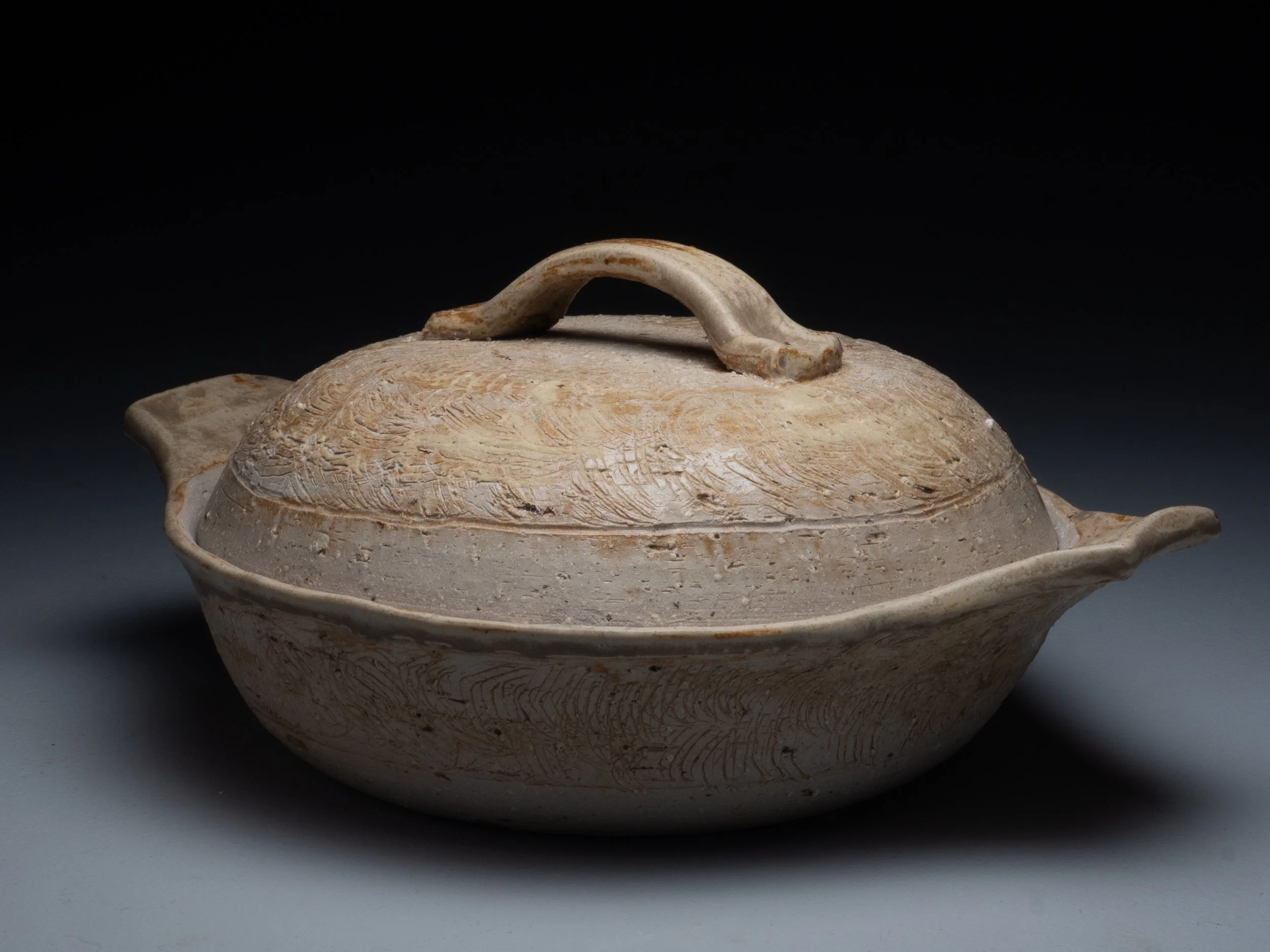 A beige ceramic teapot with a textured surface and a curved handle on the lid, placed on a light gray surface against a dark background.