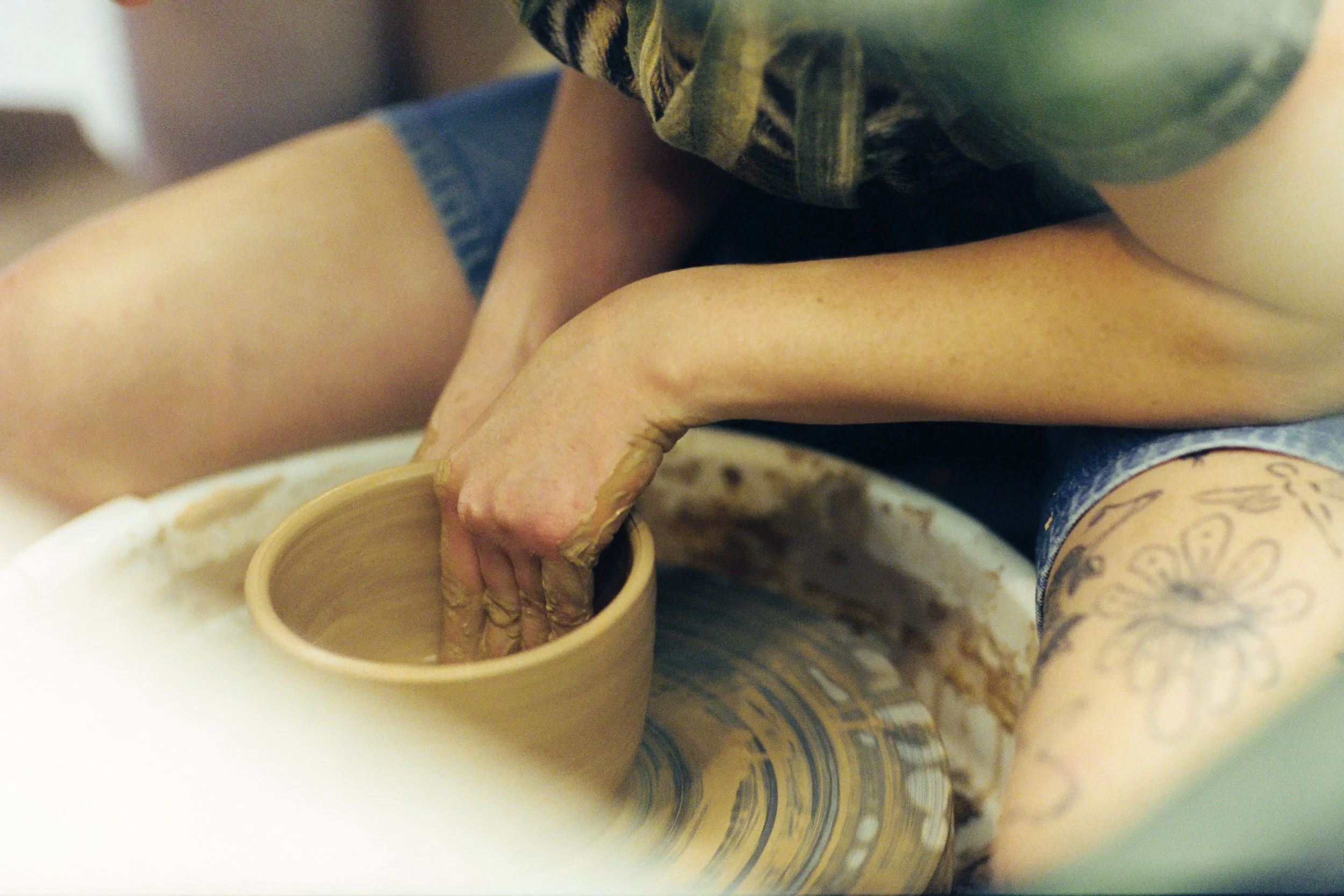 Close-up of a person shaping pottery on a pottery wheel, wearing a green shirt and blue patterned pants.
